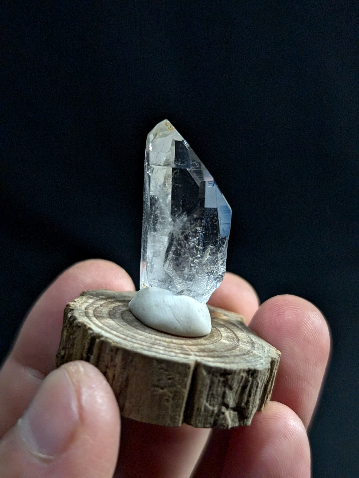 A clear, faceted quartz crystal with a pointed tip stands upright on a smooth white stone, which rests atop a rustic wooden slice held in someone’s hand.