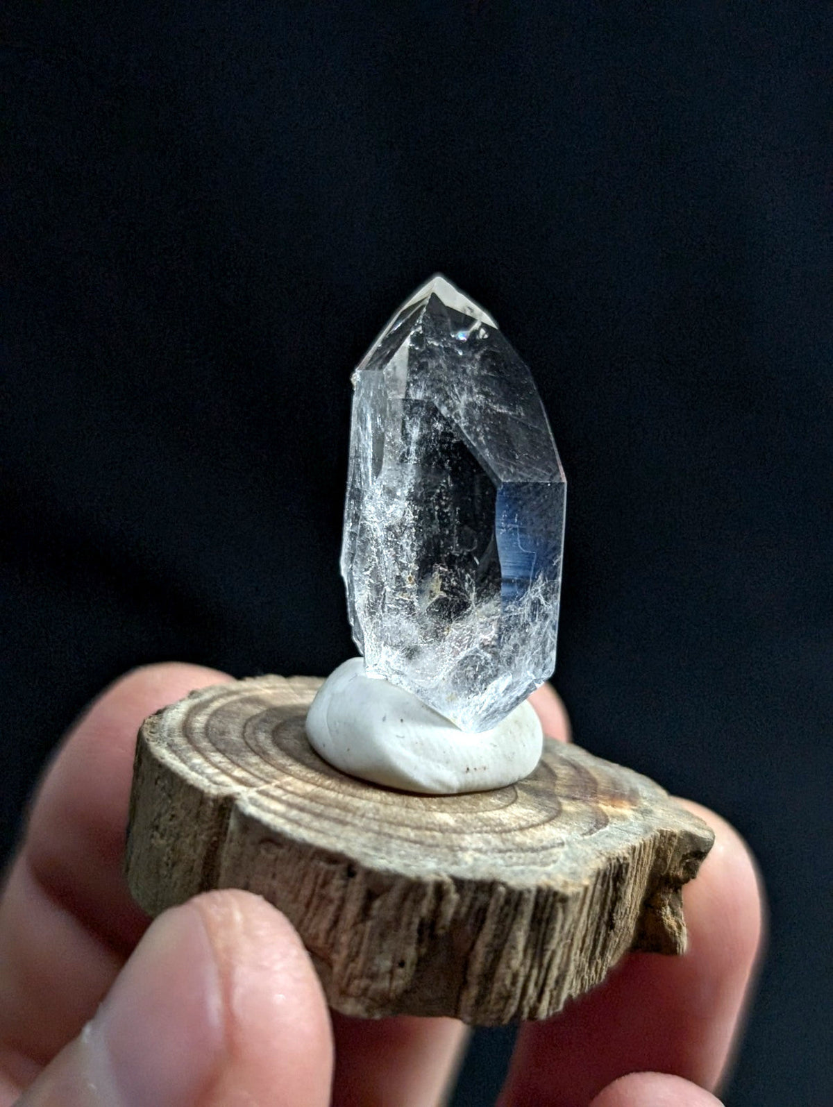 A clear, faceted quartz crystal with subtle internal inclusions sits atop a white ceramic base, which is mounted on a rustic wooden slice.