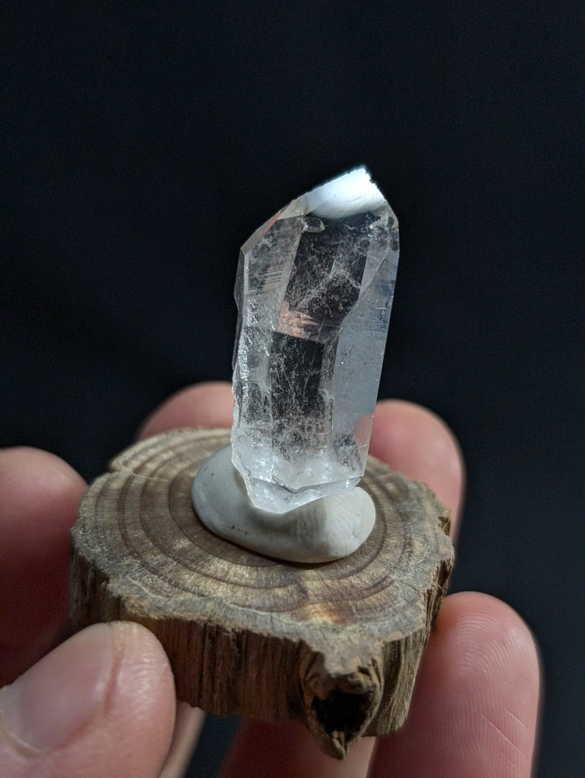 A clear, faceted quartz crystal with a pointed tip and visible internal inclusions is displayed on a rustic wooden slice base.