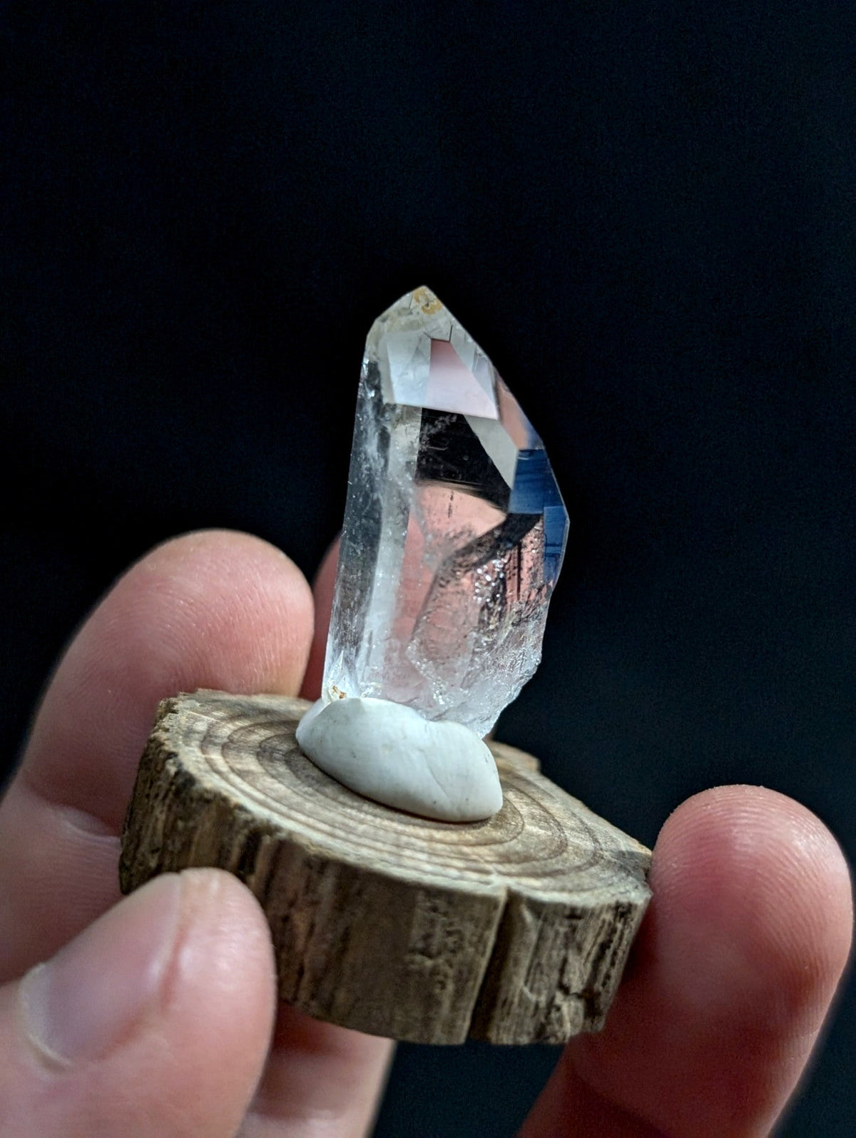 A clear quartz crystal with a pointed tip, mounted on a smooth white stone that sits atop a rustic wooden slice.