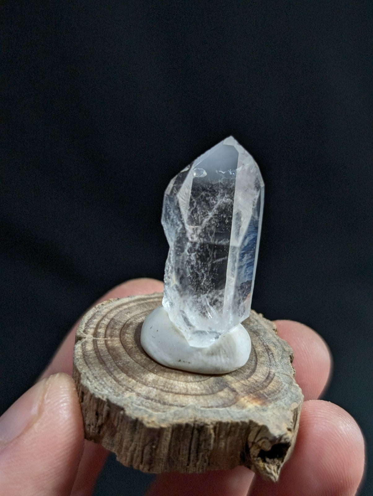A clear quartz crystal with a pointed tip and visible internal inclusions, mounted on a smooth white base that sits atop a rustic wooden slice.