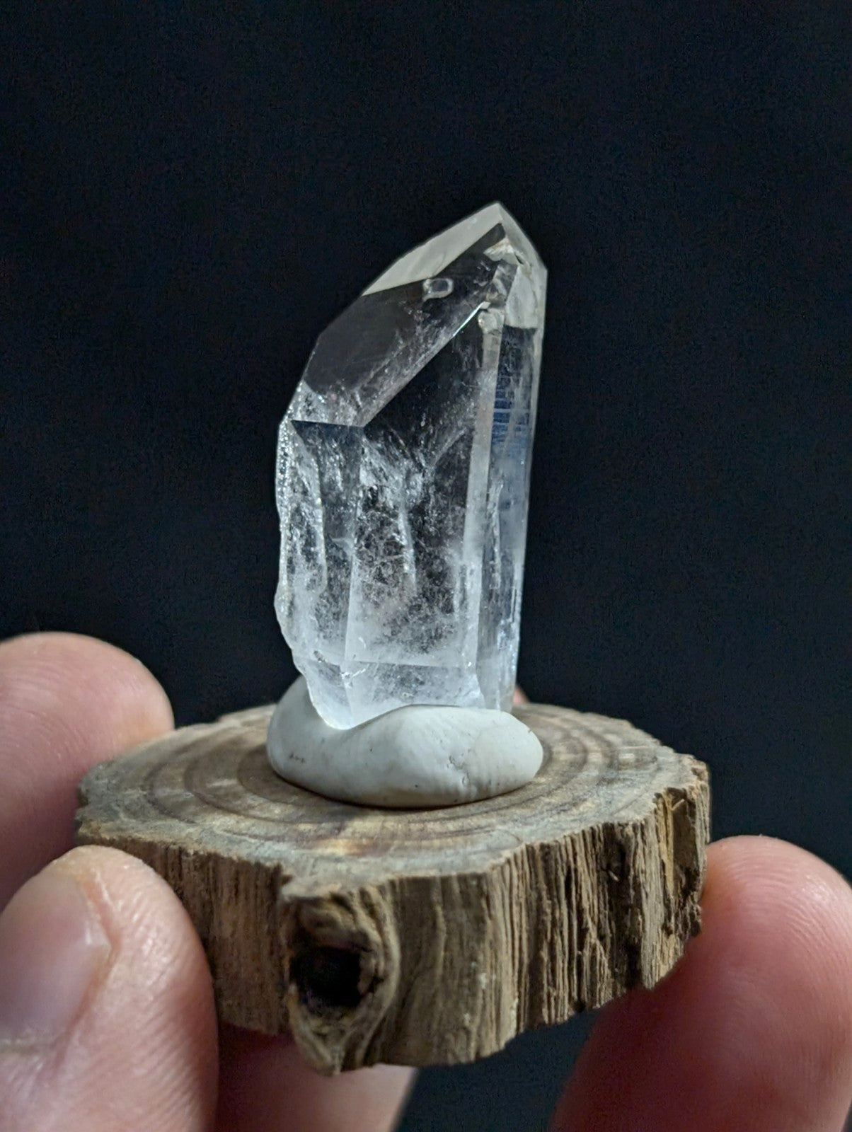 A clear, faceted quartz crystal with a pointed tip and internal inclusions sits atop a smooth white stone on a rustic wooden slice.