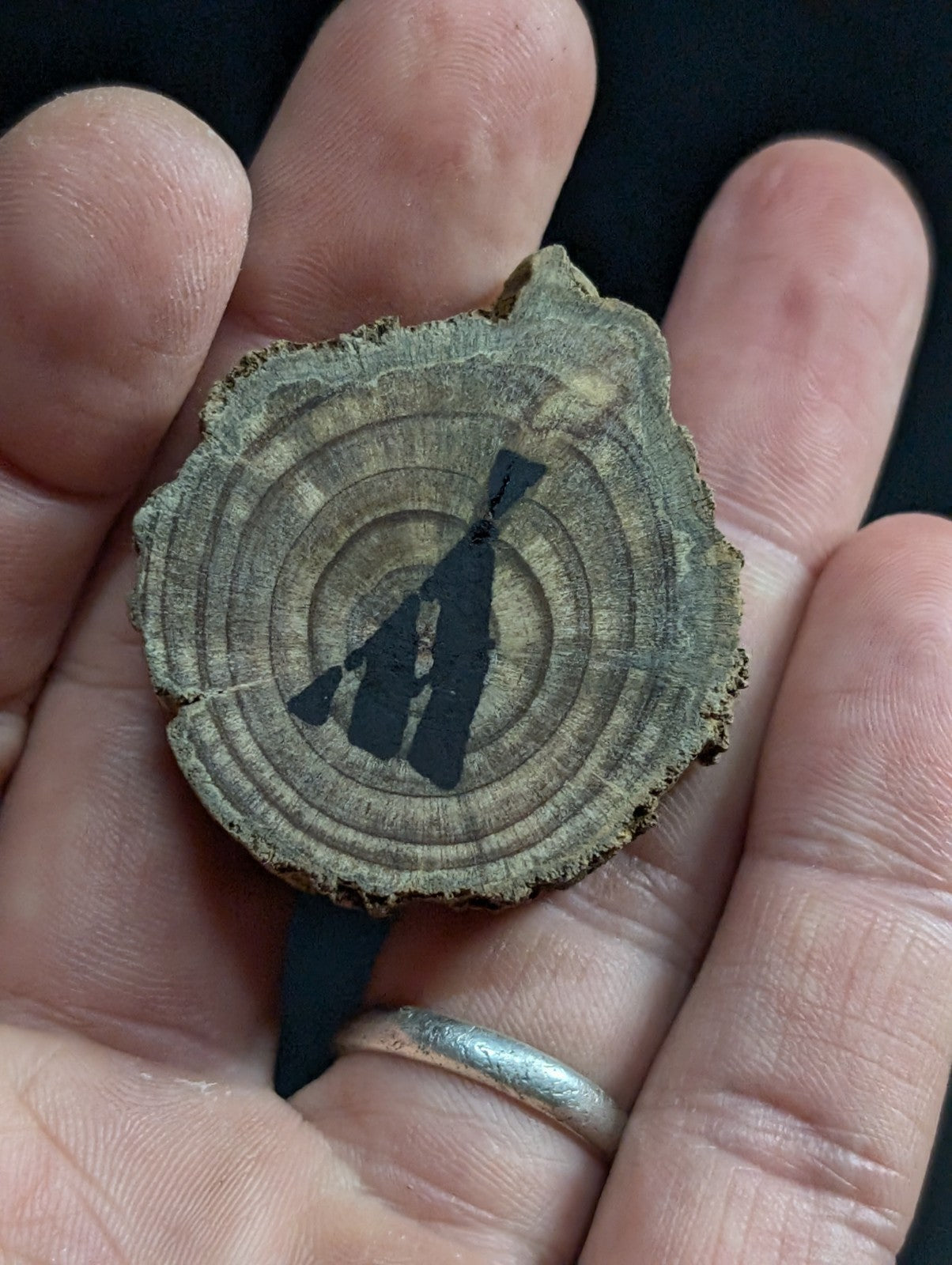 A rustic, circular wooden slice pendant featuring a black silhouette of a figure holding a rifle against its natural wood grain rings.