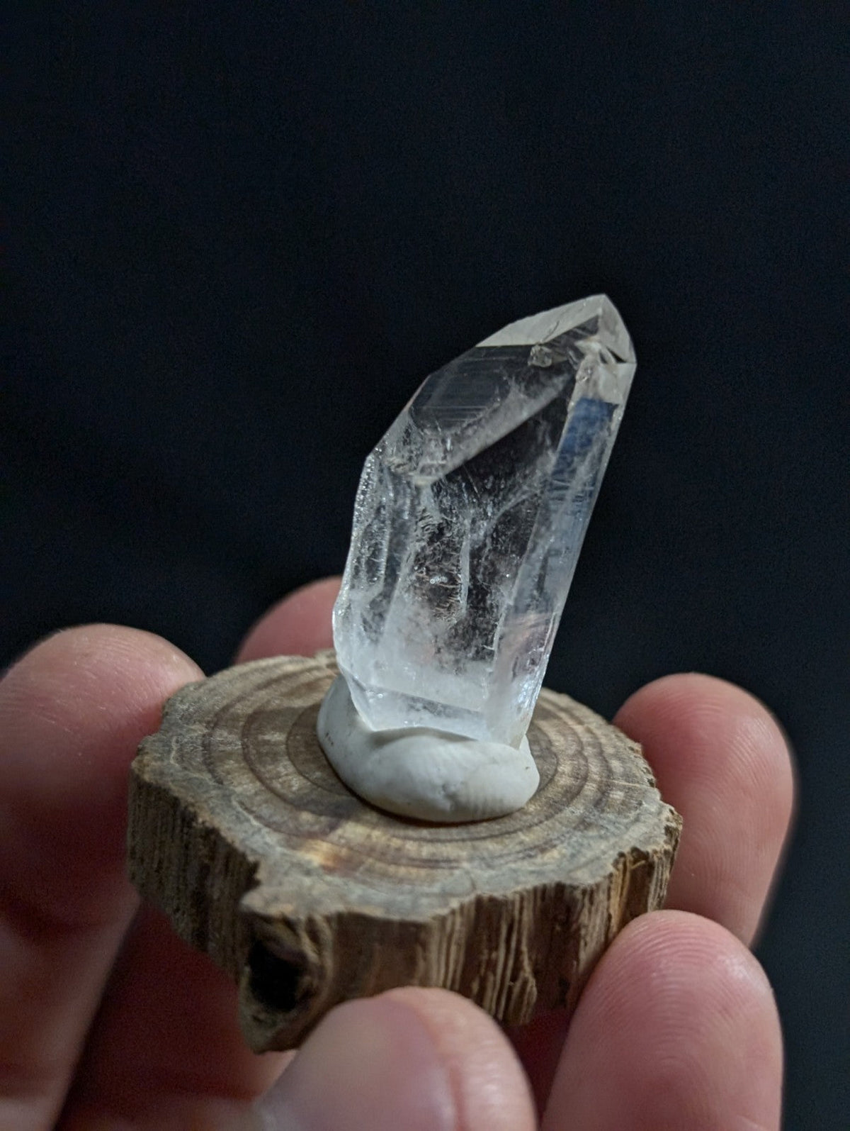 A clear, faceted quartz crystal with natural internal inclusions sits atop a rustic wooden slice.