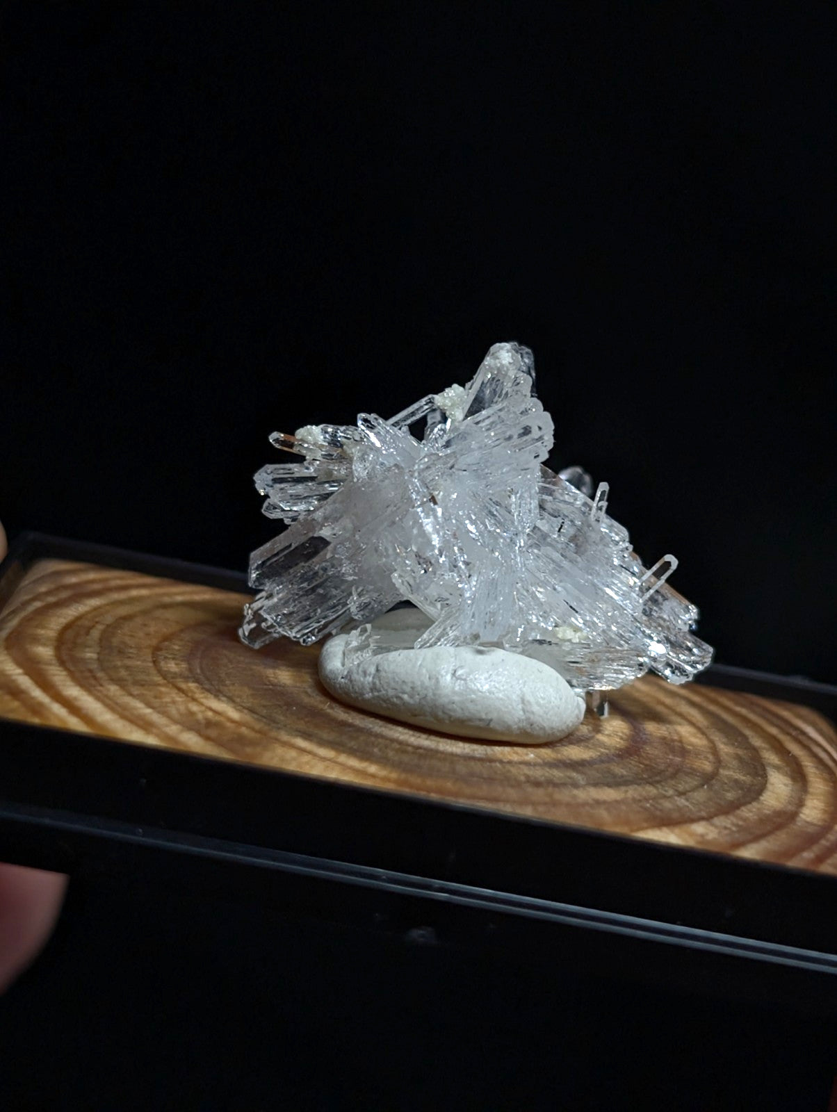 A clear, translucent quartz crystal cluster with sharp, interlocking facets rests atop a smooth white stone on a wooden display surface.