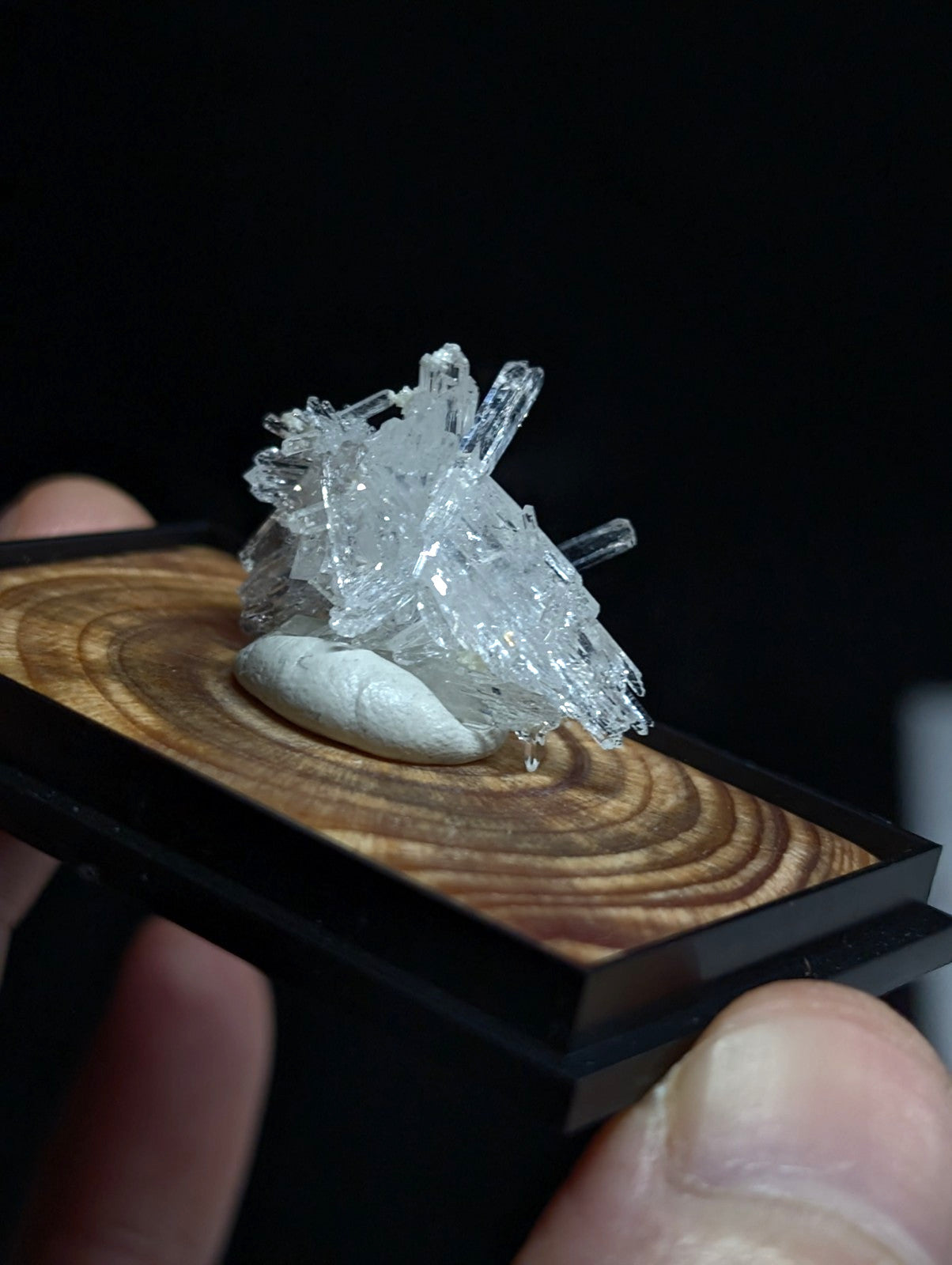 A clear, crystalline mineral specimen with sharp facets and a translucent quality rests atop a smooth white stone within a black-framed wooden display tray.