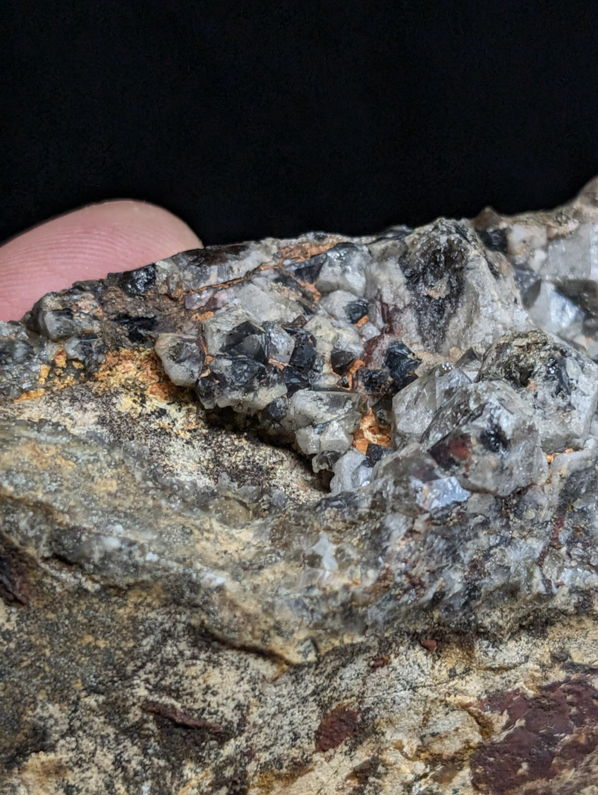 A rugged, multi-toned mineral specimen featuring a cluster of translucent gray and black crystals embedded in a rough, earthy matrix with patches of orange and reddish hues.
