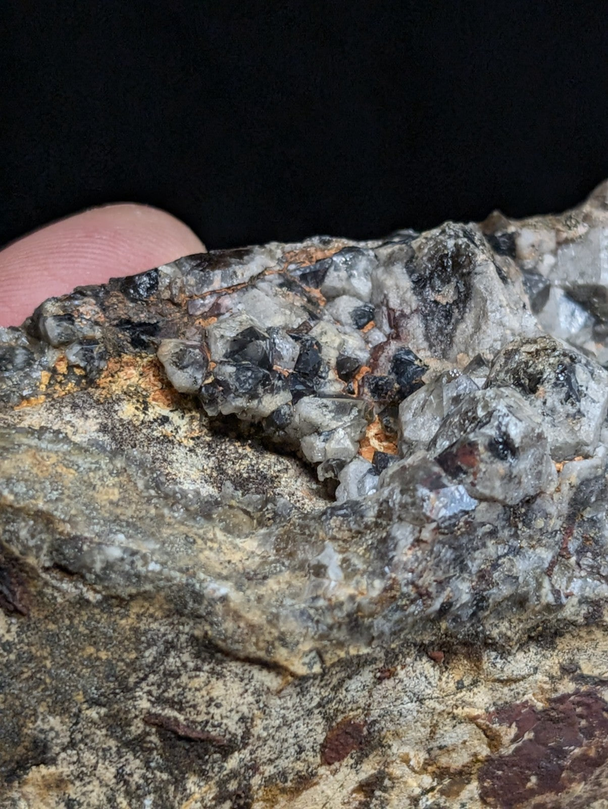 A rugged, multi-toned mineral specimen featuring a cluster of translucent gray and black crystals embedded in a rough, earthy matrix with patches of orange and reddish hues.