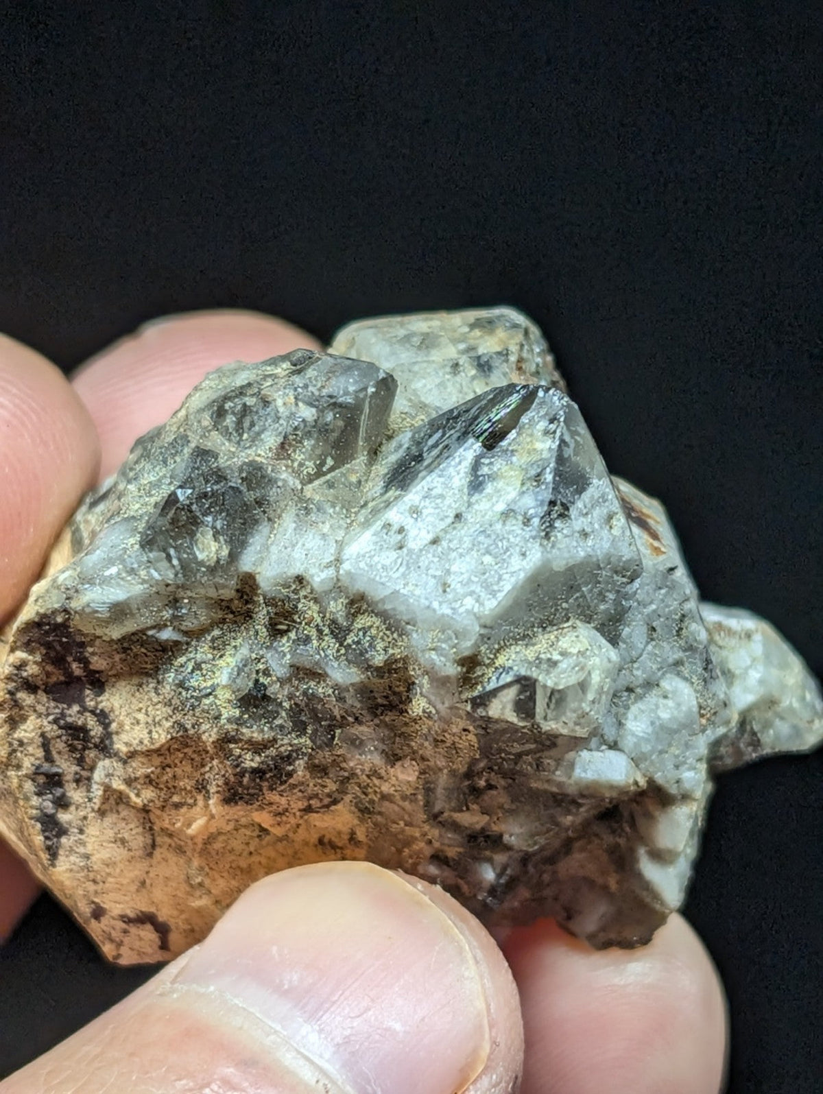 A hand holds a rough, translucent quartz crystal cluster with clear and smoky gray facets against a dark background.