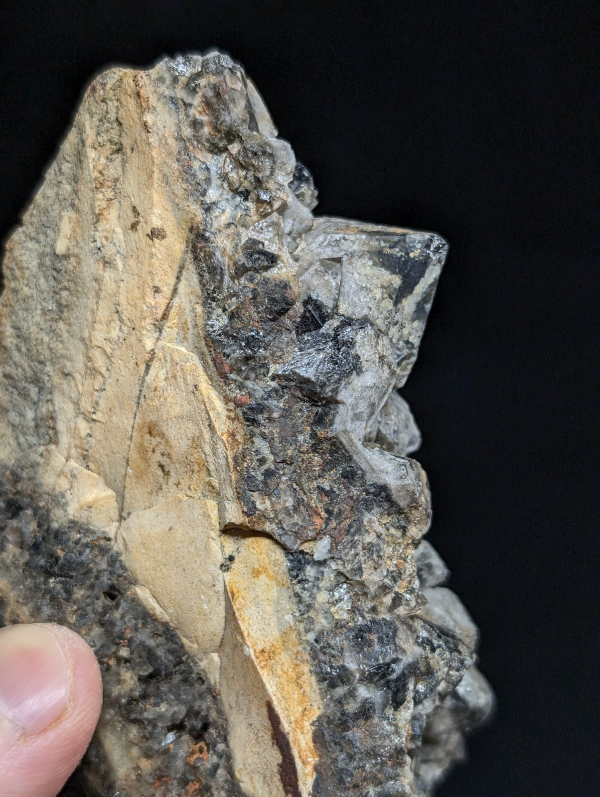 A rugged, multi-toned mineral specimen featuring translucent gray crystals embedded in a matrix of earthy beige and dark speckled rock.
