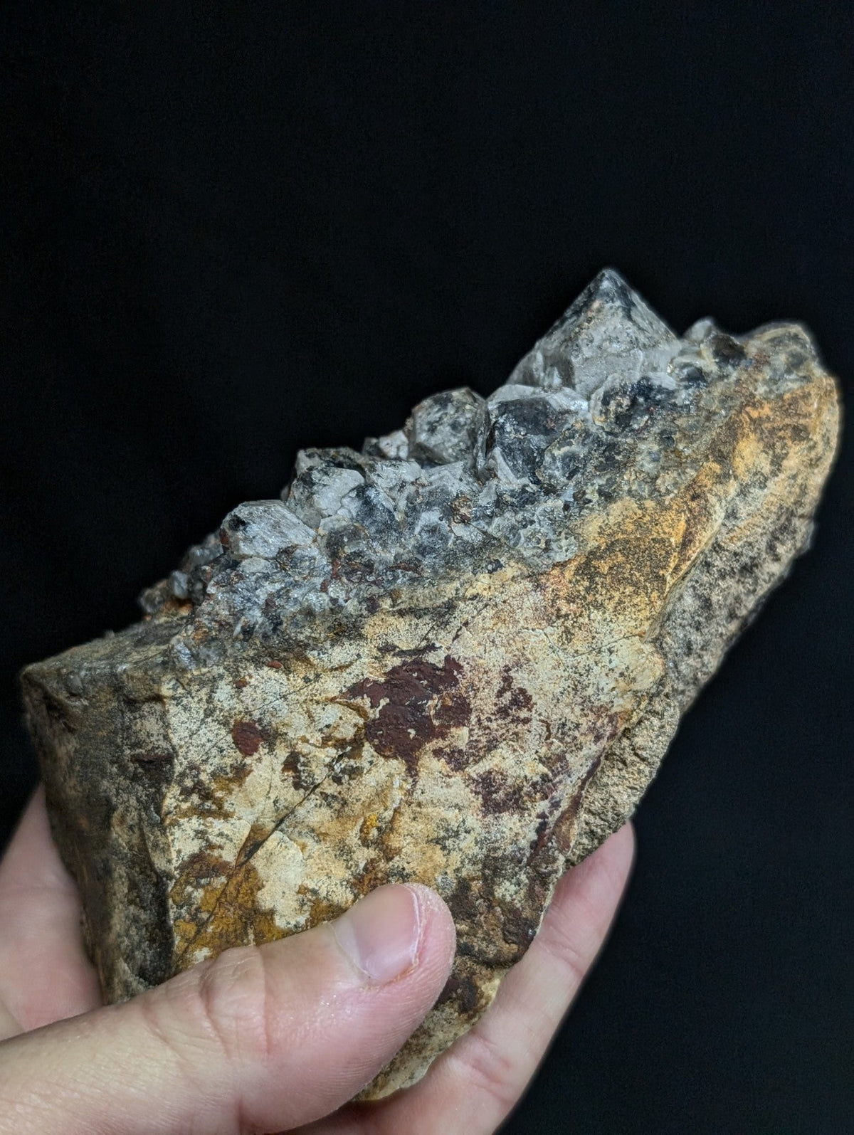 A hand holds a rough, irregularly shaped rock with a textured surface featuring patches of brown and yellowish hues, topped by a cluster of translucent gray crystals.