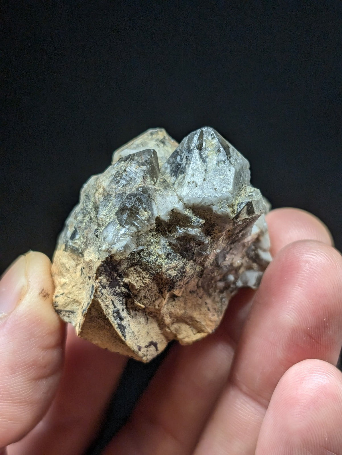 A cluster of translucent gray quartz crystals with a rough, earthy base held in someone’s hand.