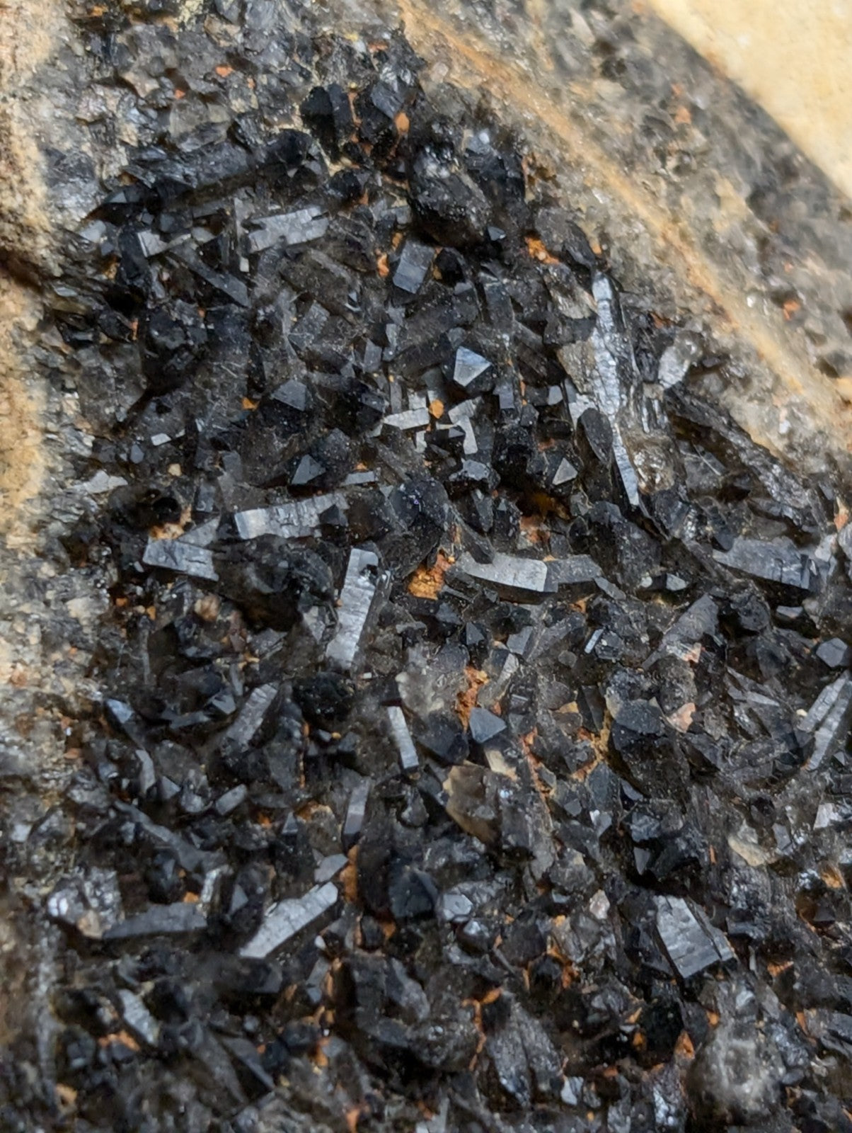 A cluster of dark, crystalline minerals with sharp, angular facets and subtle metallic sheen embedded in a rough-textured rock matrix.