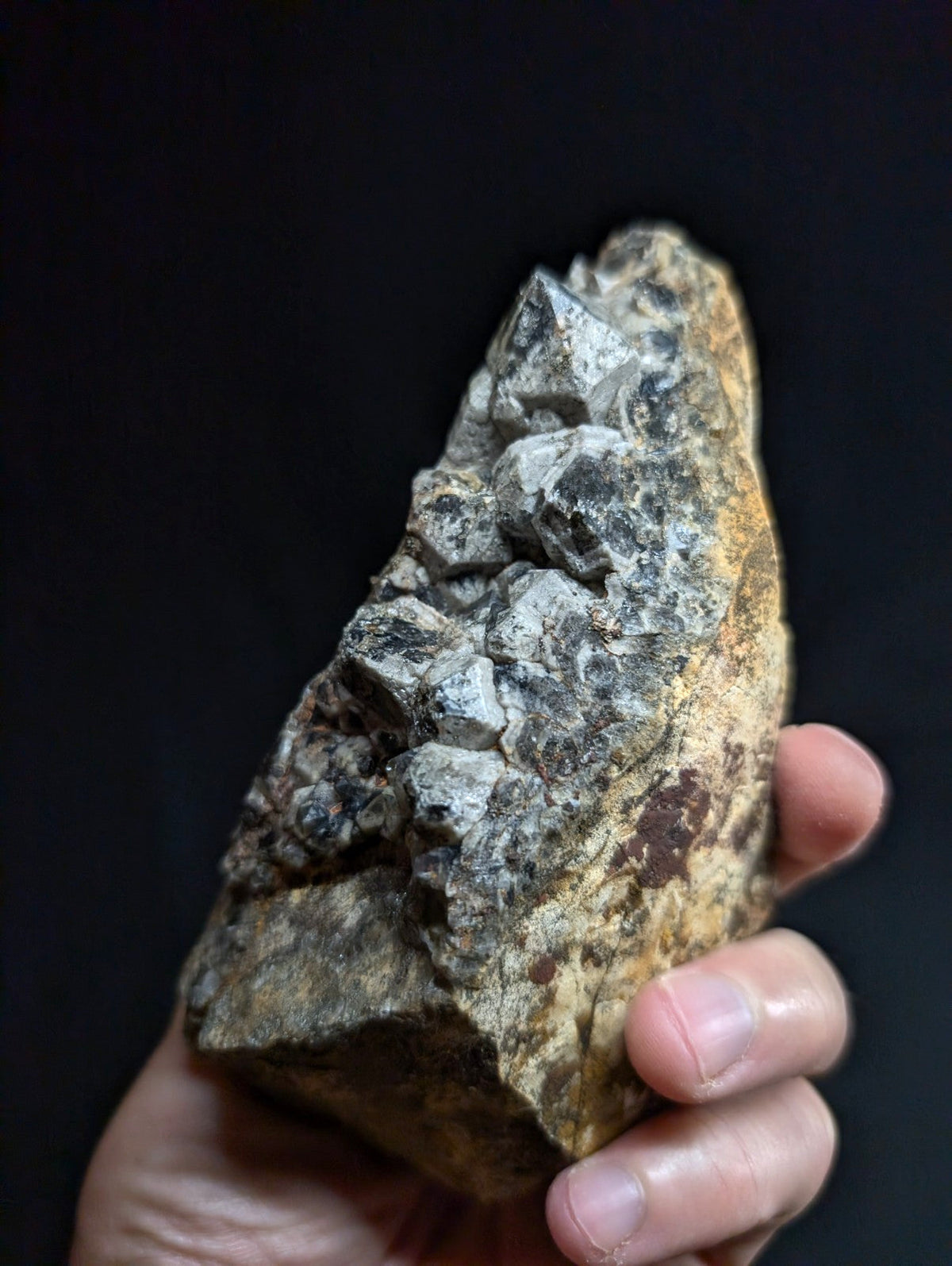 A hand holds a rough, irregularly shaped mineral specimen featuring prominent gray crystalline formations emerging from a darker, earthy matrix with patches of brown and yellow.
