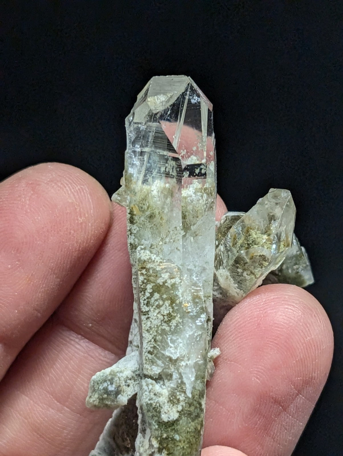 A clear quartz crystal cluster with greenish mineral inclusions, held gently between fingers.