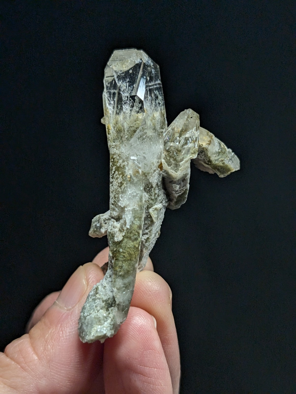 A translucent, clear quartz crystal formation with a rough, earthy greenish-gray matrix holding together multiple pointed crystalline structures.