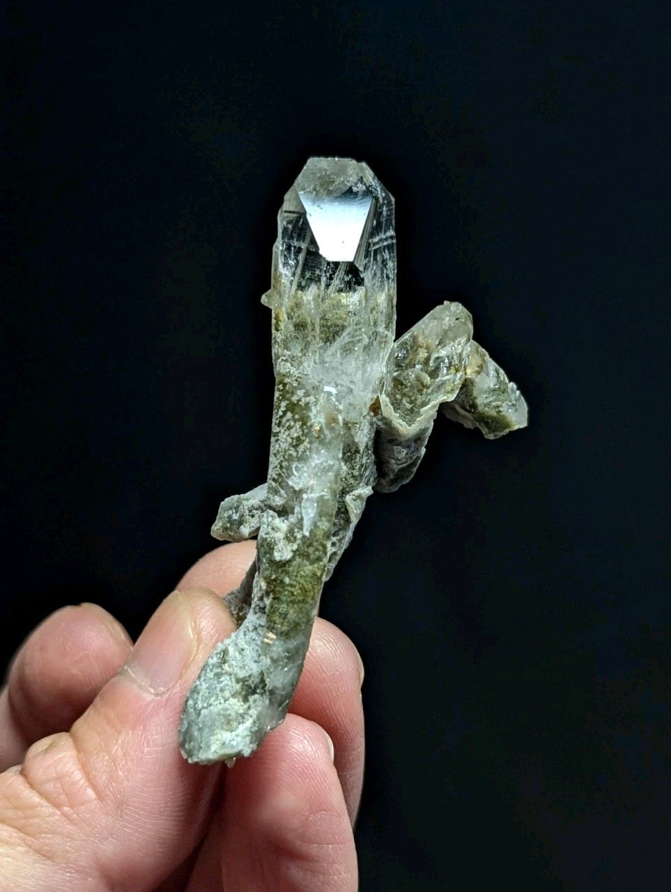 A translucent green quartz crystal with a pointed, faceted tip and rougher, mossy base held in hand.