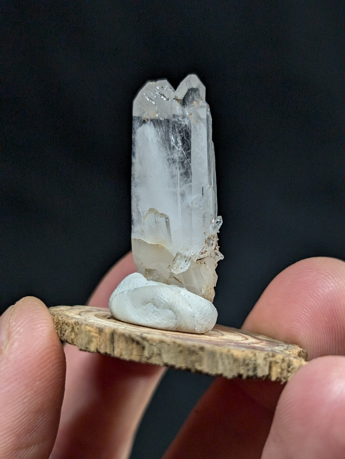 A clear, translucent quartz crystal with a pointed top and natural facets stands upright on a smooth white stone, which rests upon a rustic wooden slice held in someone’s fingers.