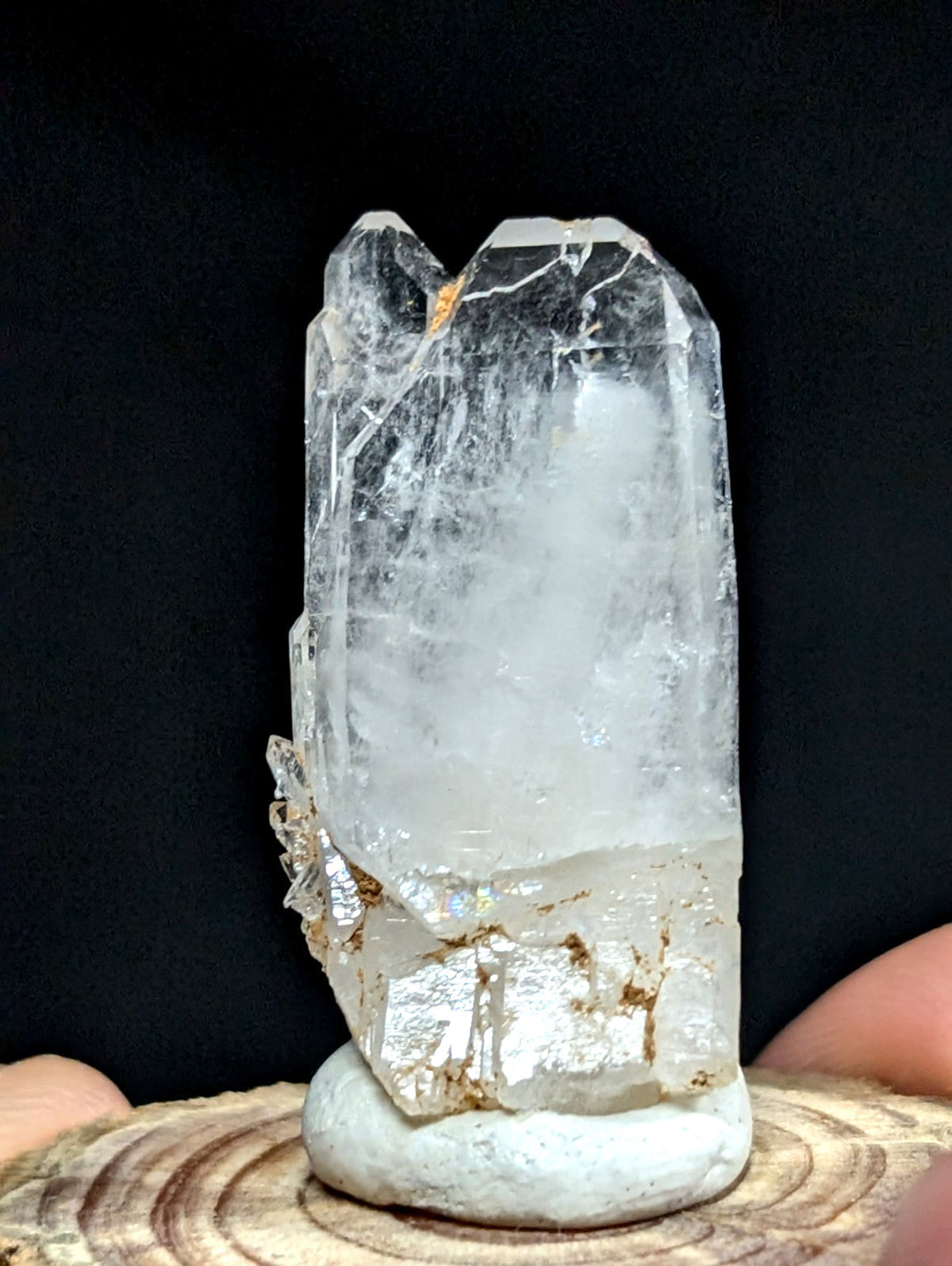 A clear quartz crystal with natural inclusions and a rough, earthy base rests on a circular white pedestal.