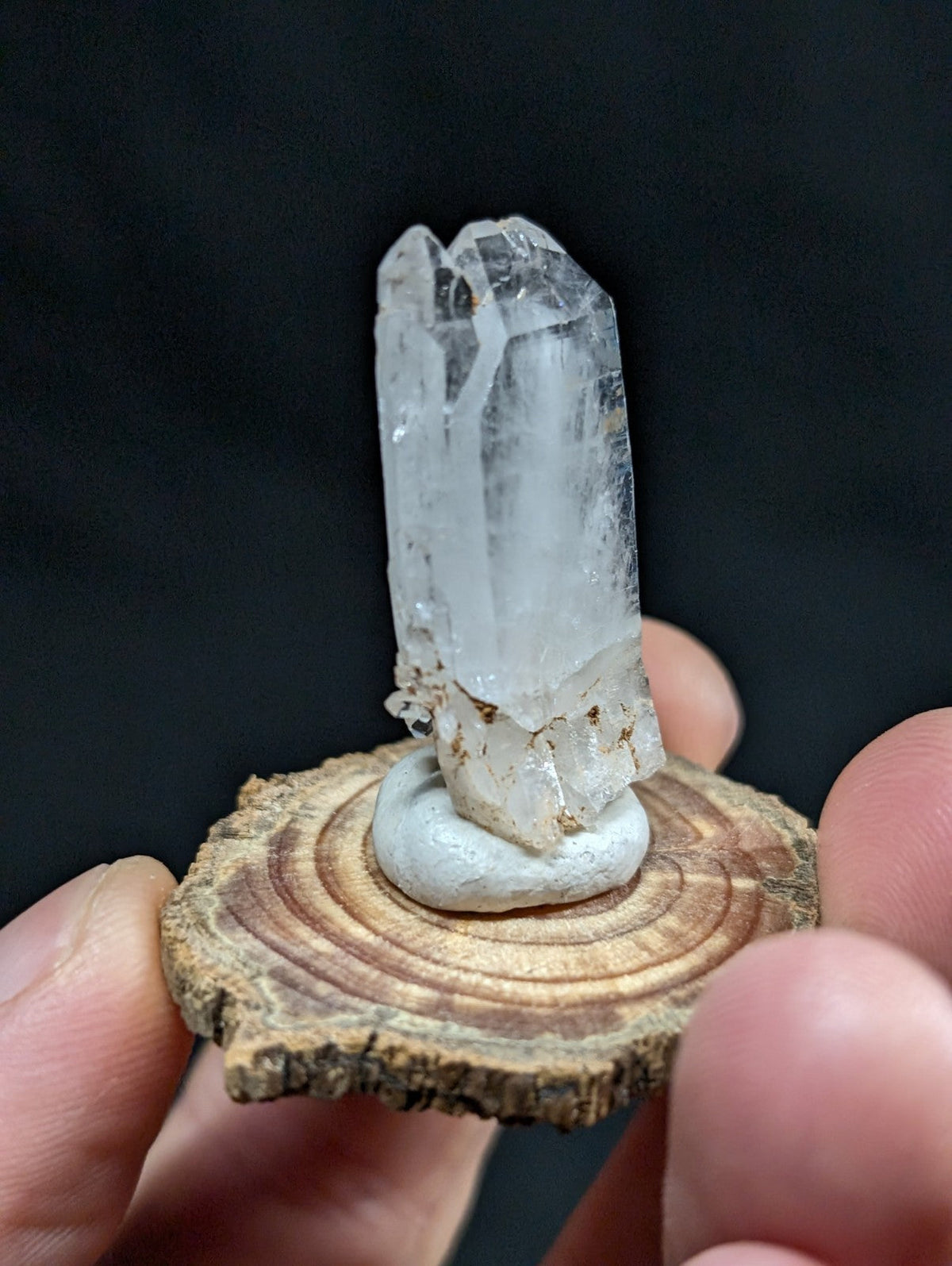 A clear quartz crystal with a faceted, translucent body and natural imperfections stands upright on a circular wooden slice base.