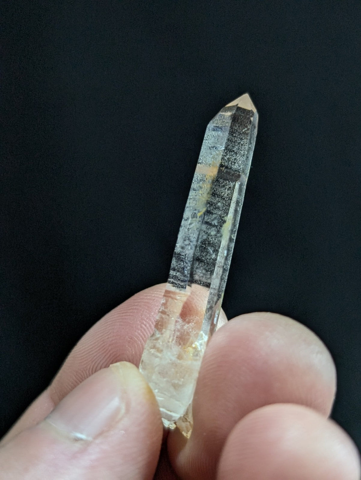 A clear quartz crystal with a pointed tip and natural inclusions, held gently between fingers against a dark background.