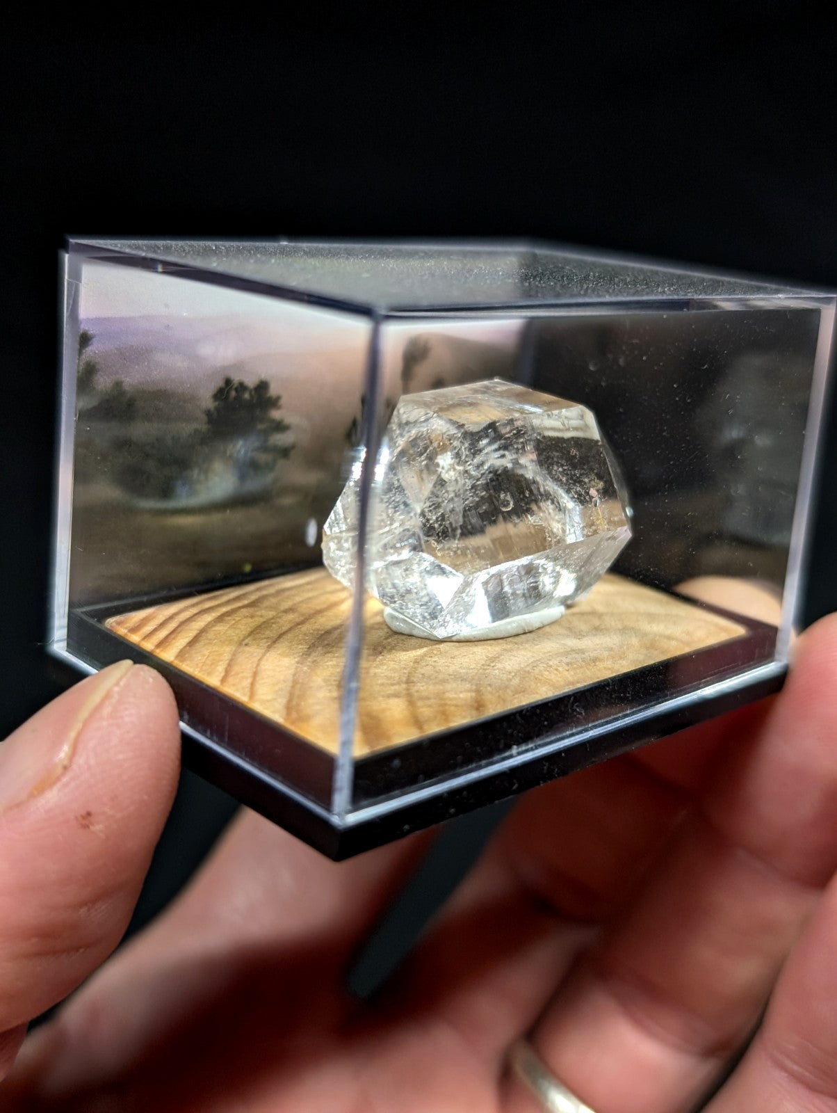 A clear, faceted quartz crystal displayed in a transparent acrylic box with a wooden base.