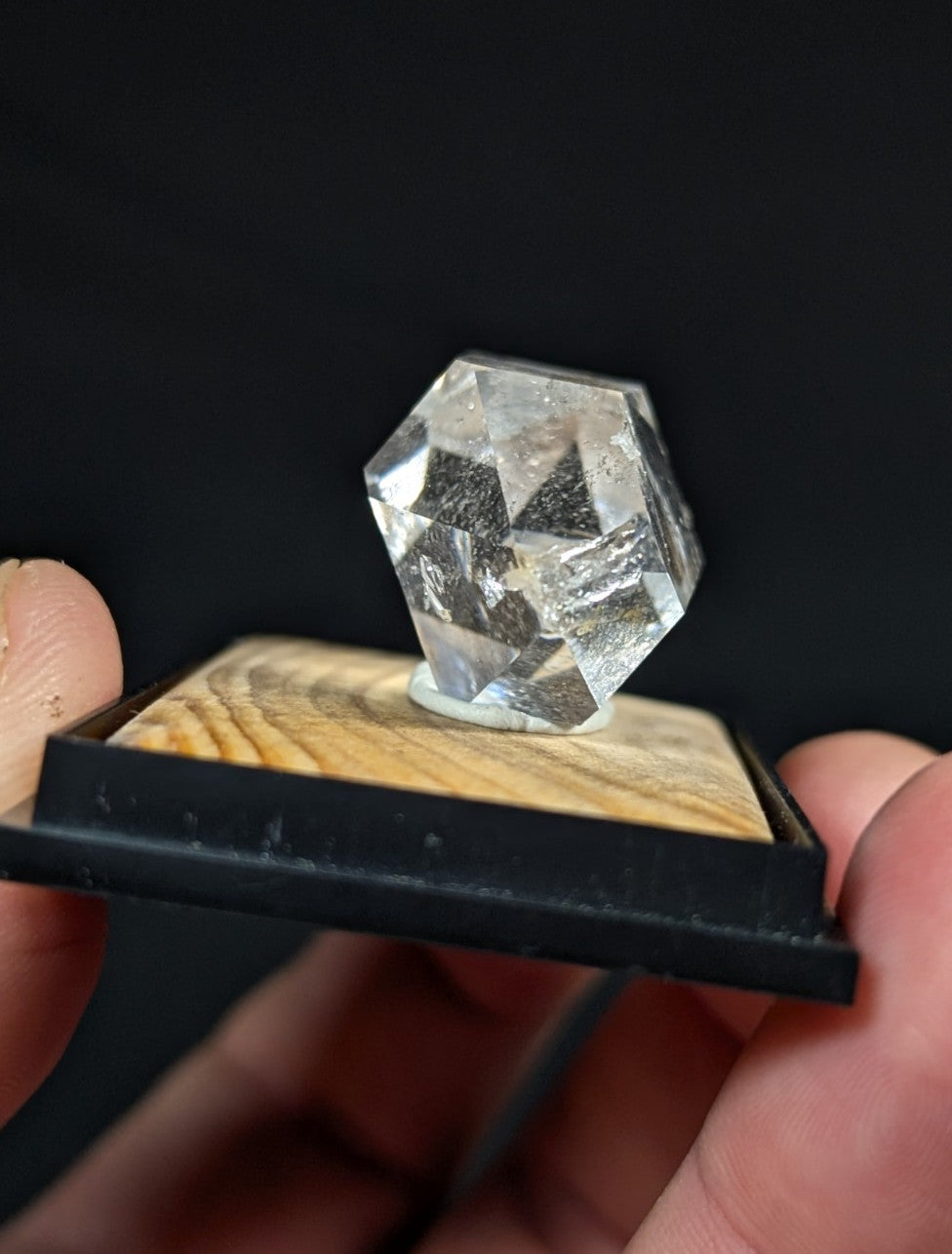 A clear, faceted quartz crystal with a hexagonal shape sits atop a wooden base encased in black plastic.