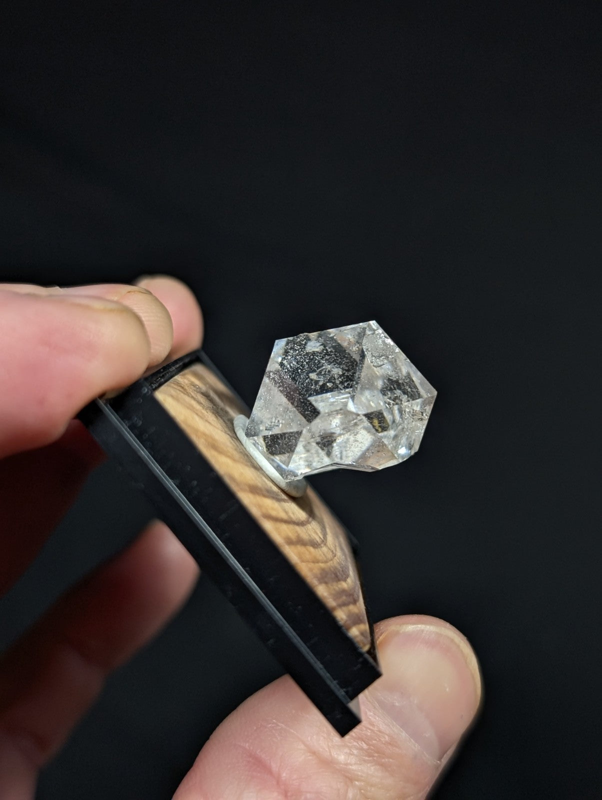 A clear, faceted quartz crystal with a hexagonal shape and natural inclusions is displayed on a wooden base within a black protective holder.