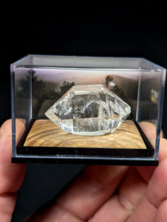 A clear, faceted quartz crystal with natural internal inclusions and a polished surface rests inside a transparent display case.