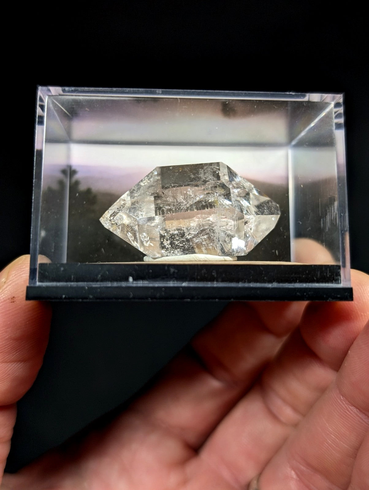 A clear, faceted quartz crystal with natural inclusions and a pointed top sits inside a transparent display case.