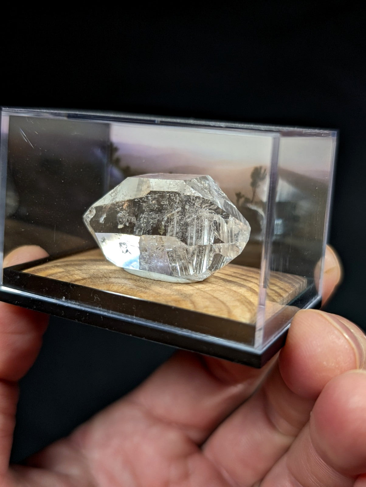 A clear, faceted quartz crystal with natural inclusions and a polished surface is displayed inside a transparent acrylic display case.