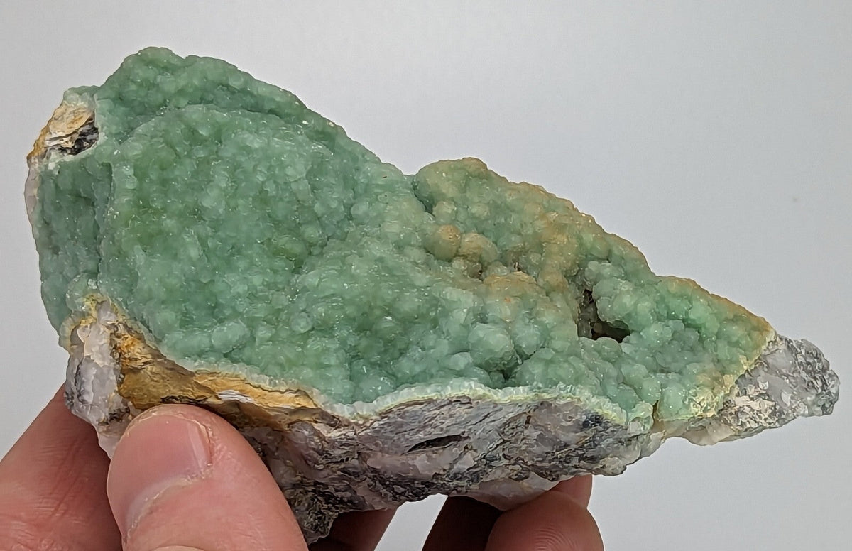 A hand holds a natural green mineral specimen with a textured, crystalline surface and earthy yellowish-brown edges.