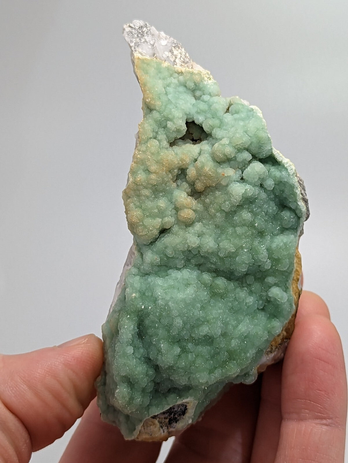 A hand holds a light green, bumpy mineral specimen with crystalline formations and patches of white and yellowish hues.