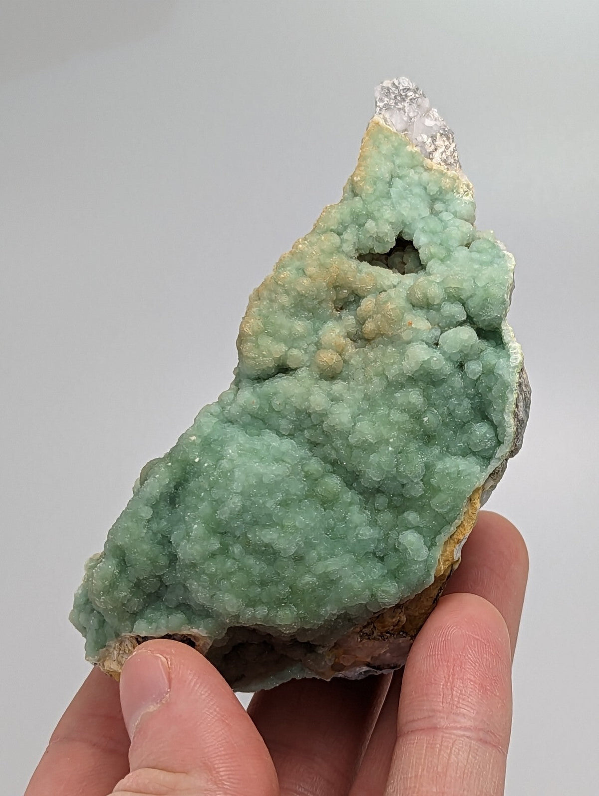 A hand holds a light green, textured mineral specimen with crystalline formations and a small clear crystal at its tip.