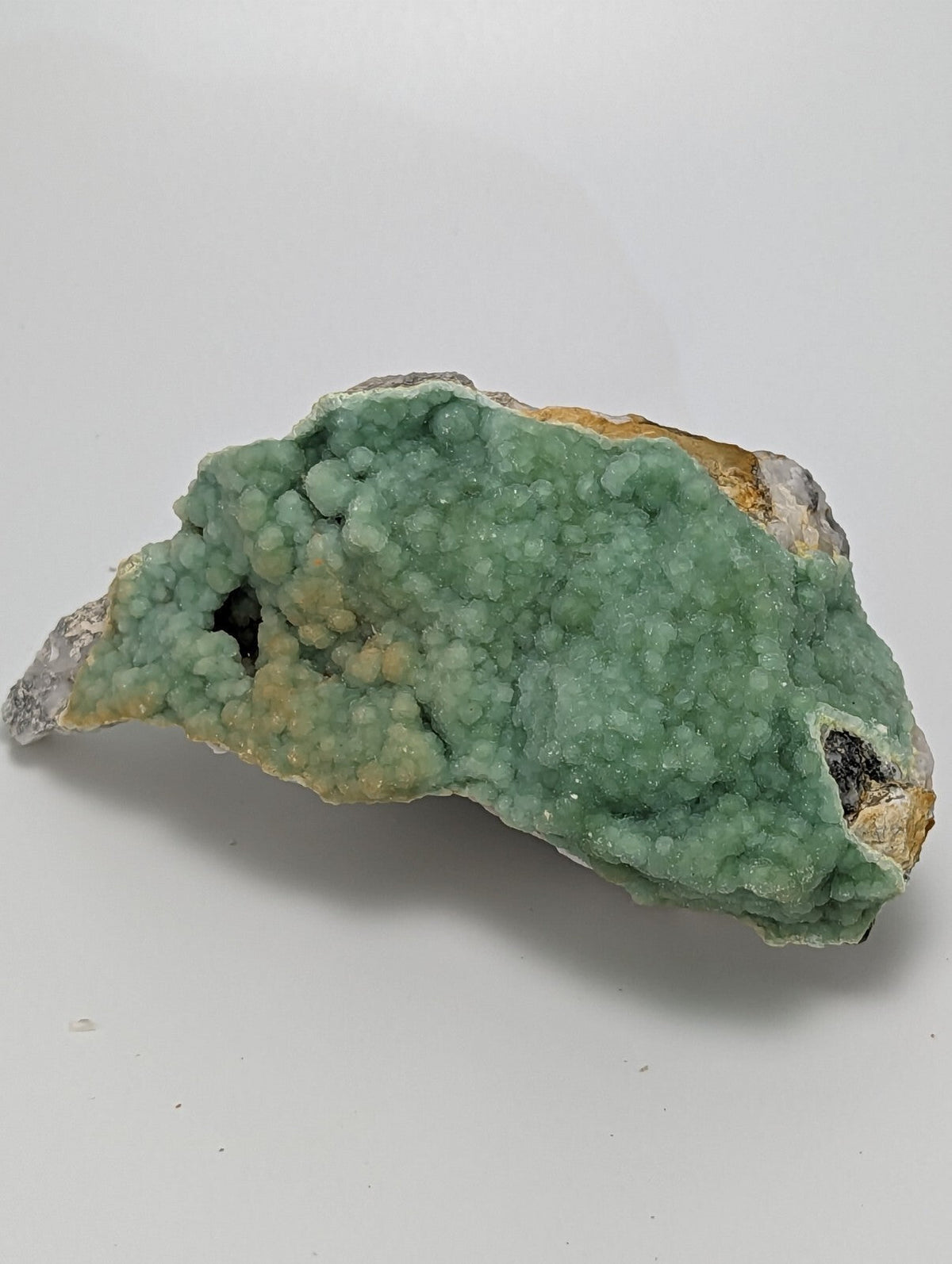 A green, crystalline mineral specimen with a textured surface and patches of yellowish and grayish material.