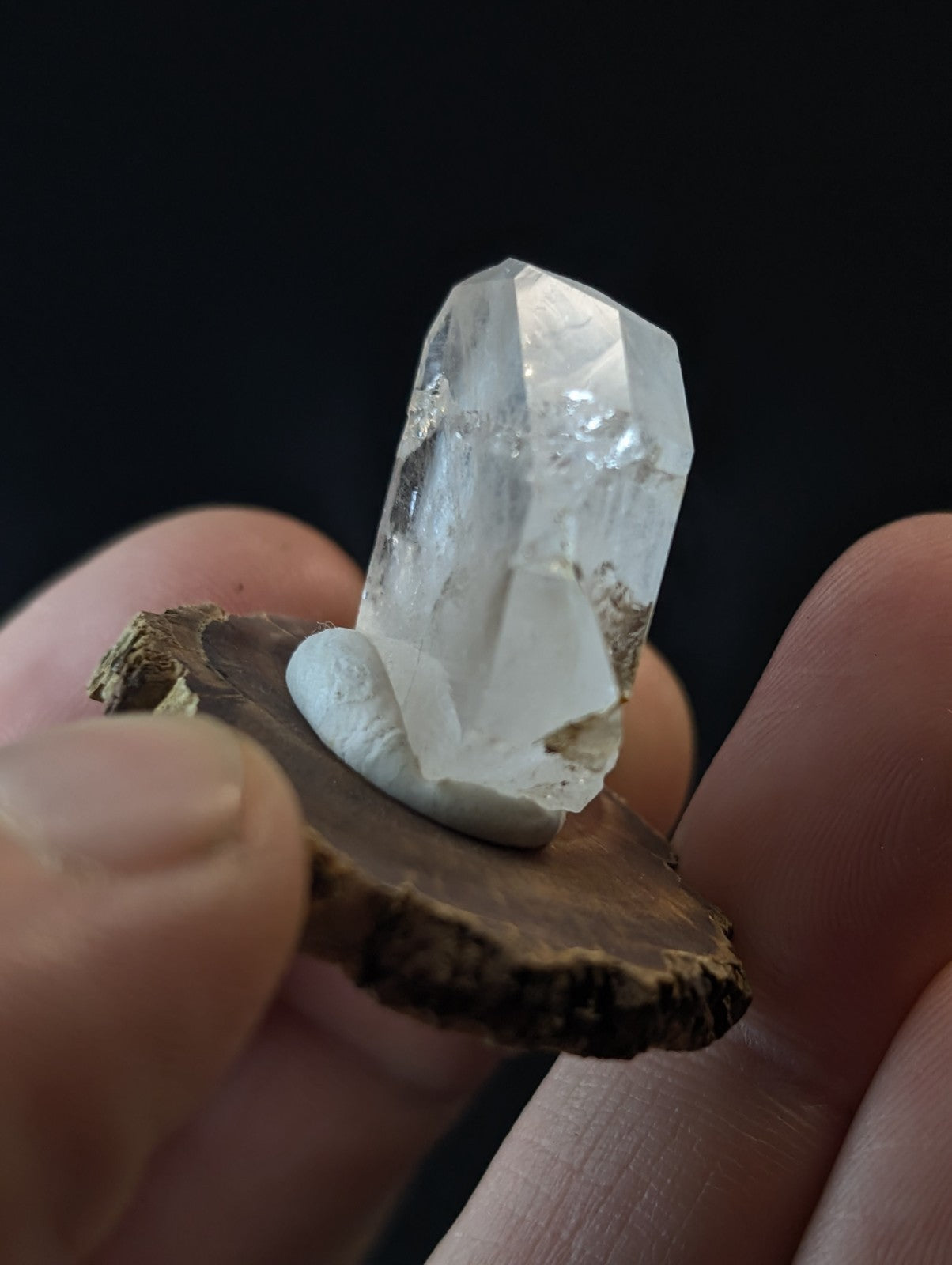 A clear, faceted quartz crystal with a rough white base is held in hand against a dark background.