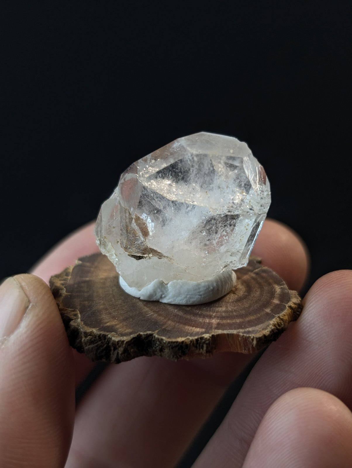 A clear, faceted quartz crystal with a natural, rough-edged form rests on a small wooden slice held gently in someone’s hand.