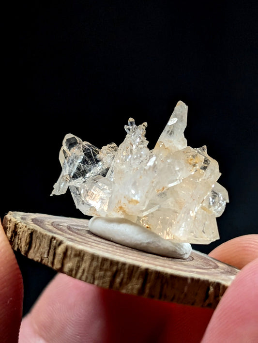 A translucent, crystalline quartz cluster with sharp facets and subtle golden inclusions rests on a smooth white stone atop a rustic wooden slice.