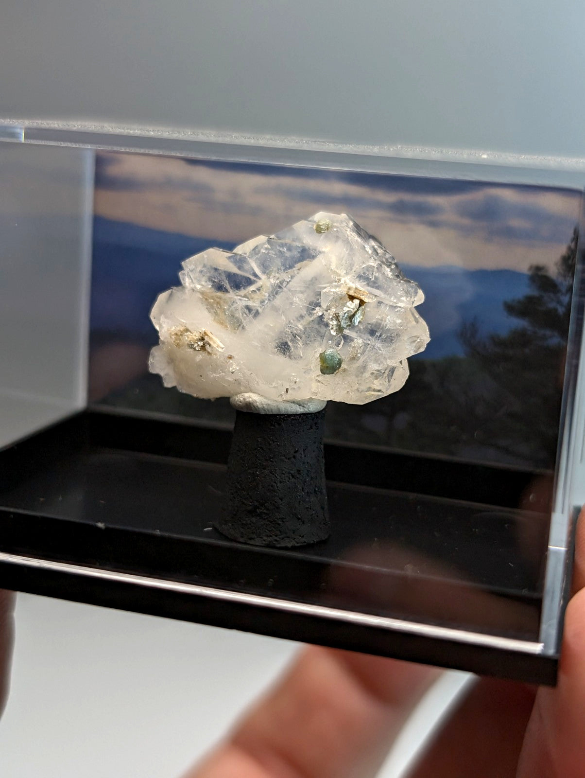 A faden quartz crystal with uv reactive blue-green cookeite and gold inclusions on black ceramic base in display case