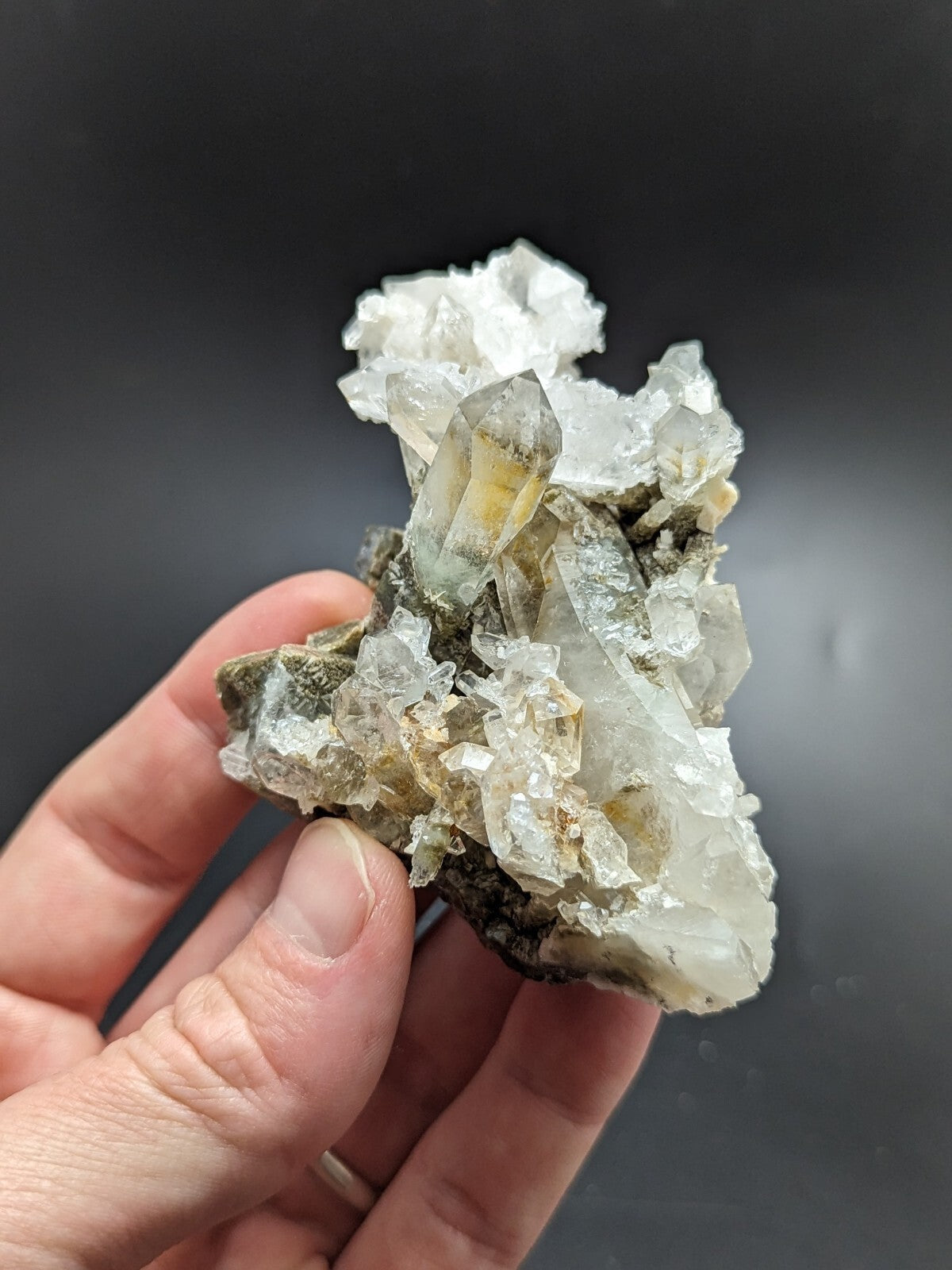 A hand holds a cluster of translucent quartz crystals with clear, pointed facets and subtle yellowish inclusions set against dark rock.