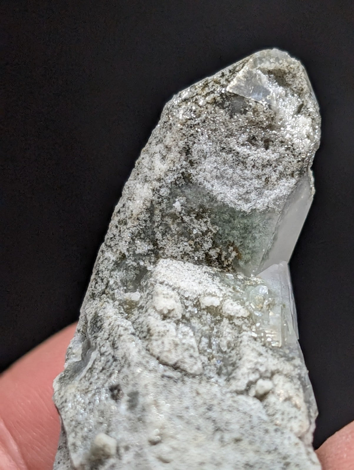 A translucent, pale green crystal with a rough, textured surface and embedded white mineral deposits.