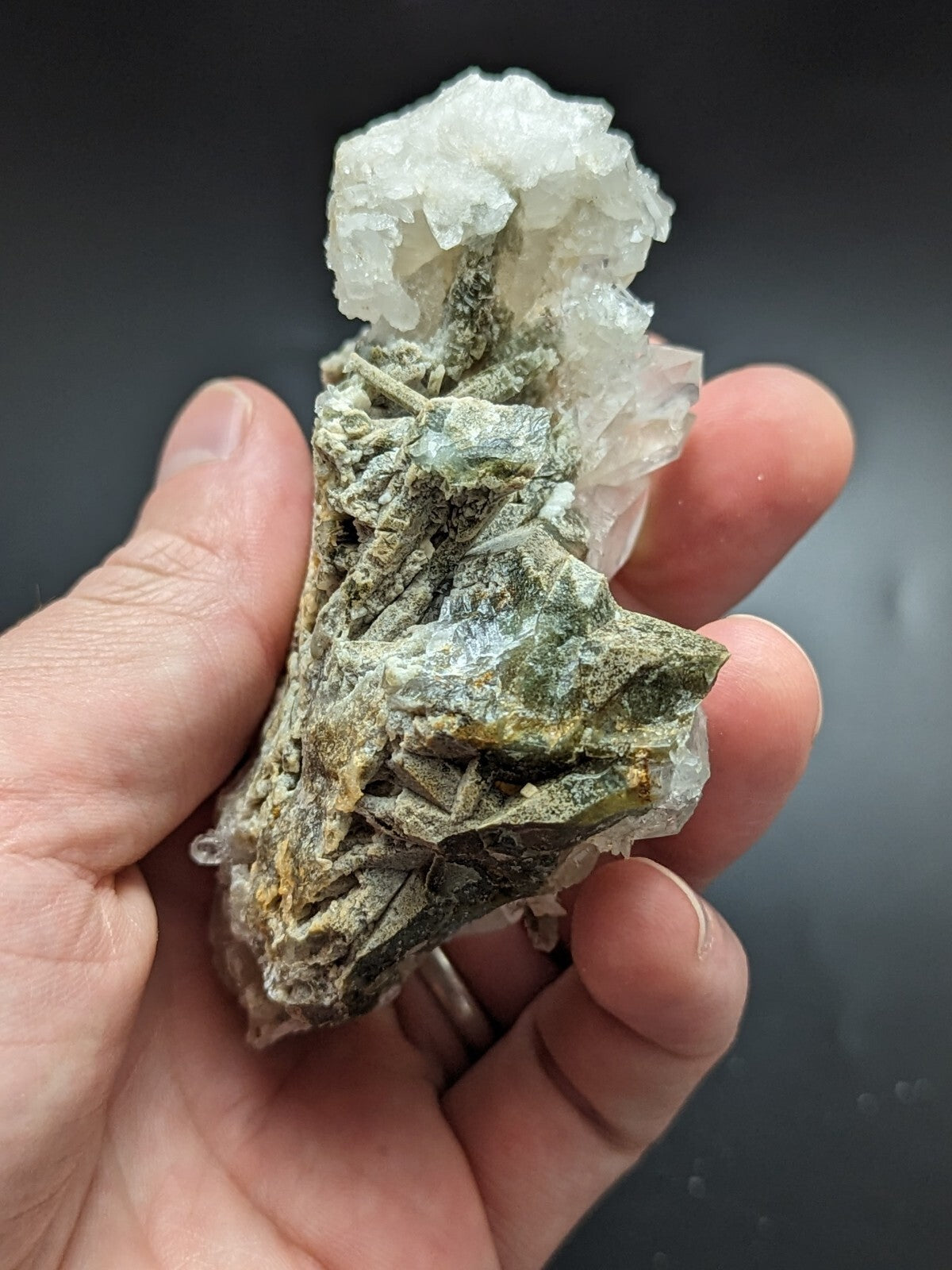 A hand holds a geological specimen featuring translucent white quartz crystals growing from a textured, greenish-gray matrix with hints of yellow.