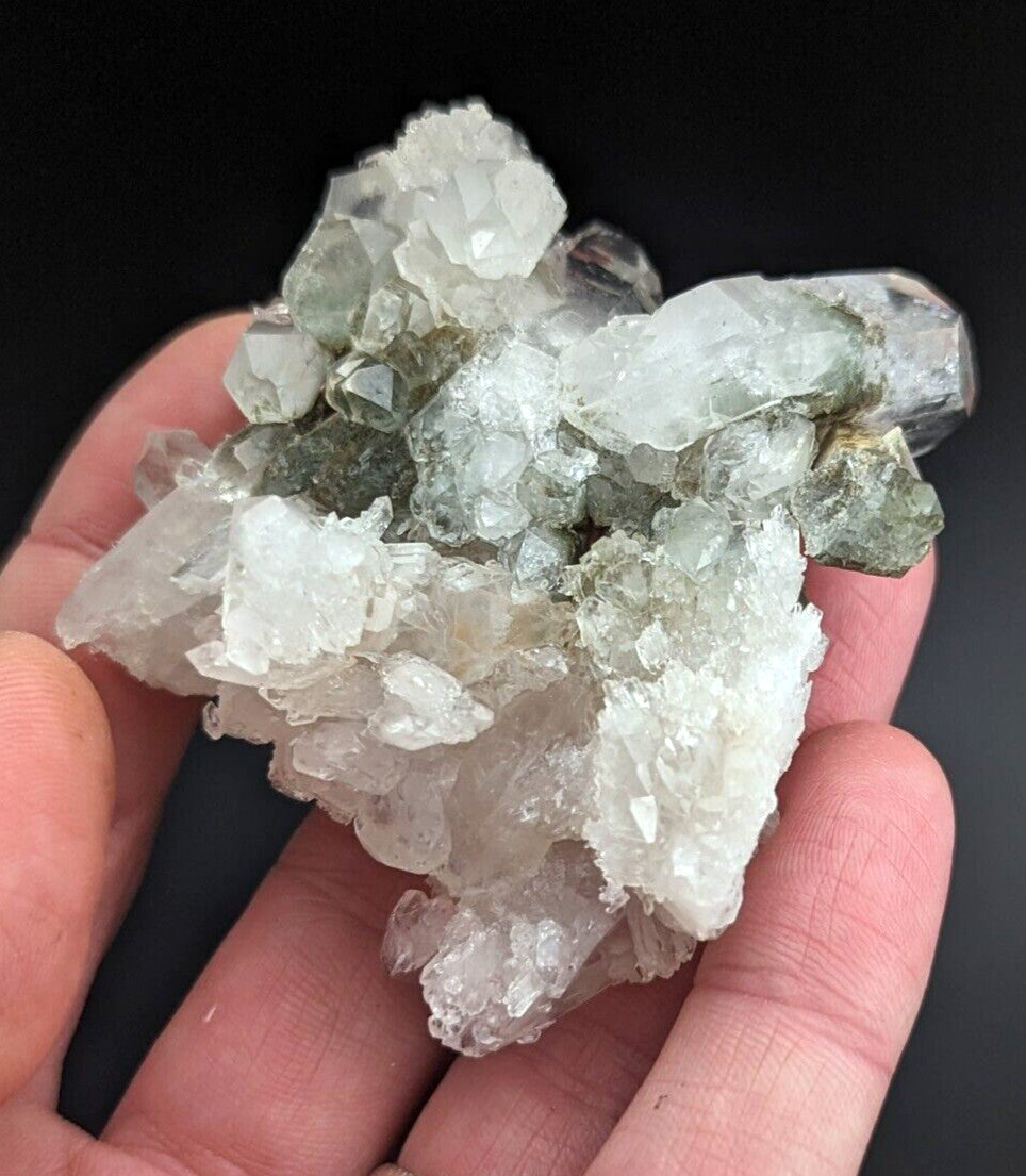 A hand holds a cluster of translucent green and clear quartz crystals with sharp, faceted edges.