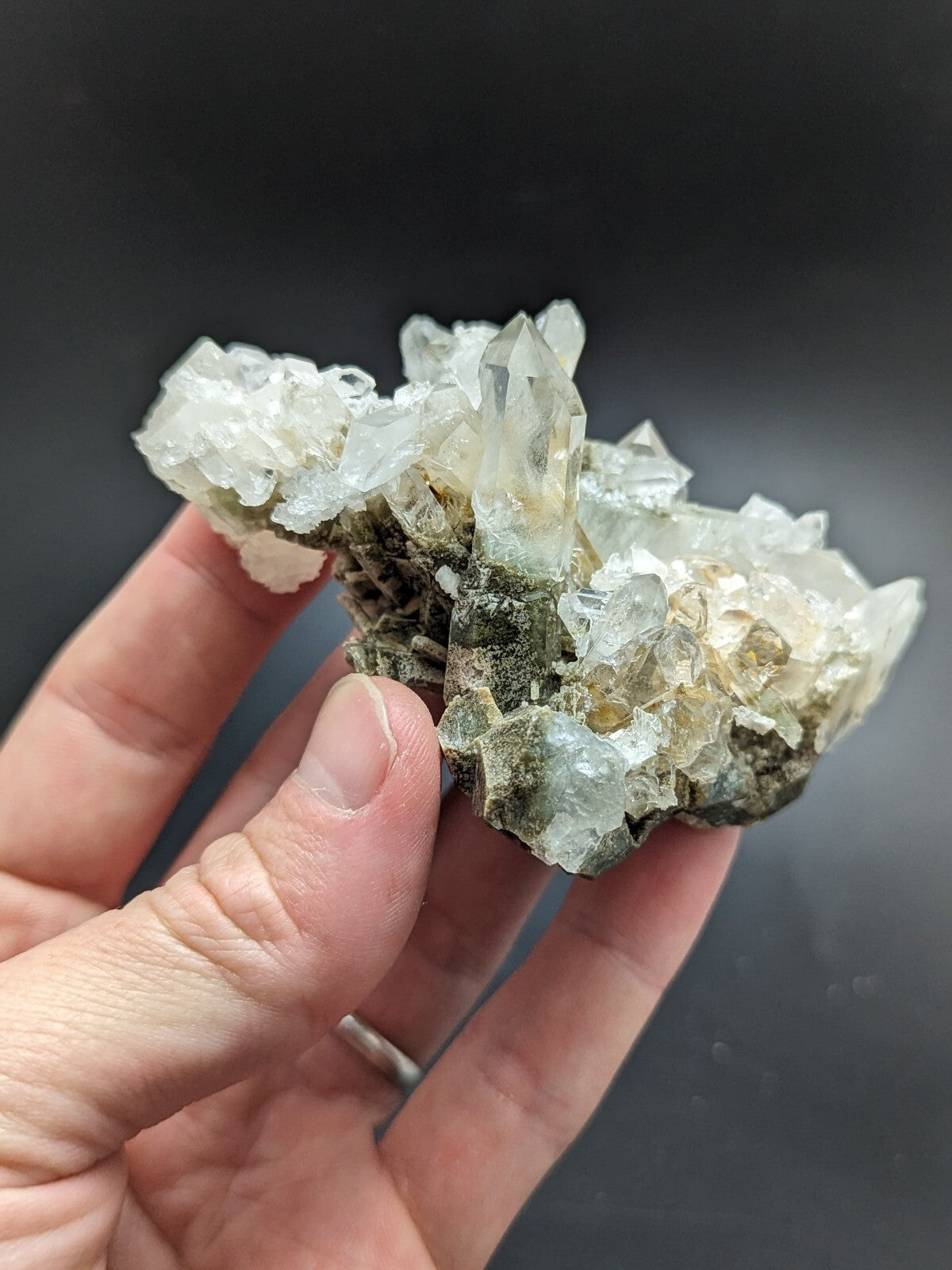 A translucent, crystalline quartz cluster with sharp, pointed facets and a mix of clear and pale yellowish hues, growing from a dark greenish-gray matrix.