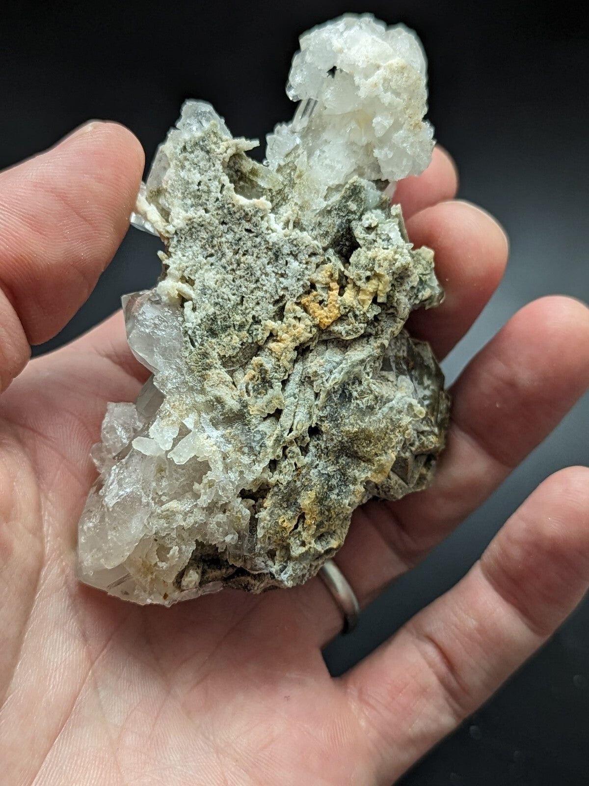 A hand holds a natural mineral specimen featuring translucent white crystals with greenish-gray matrix and subtle yellow inclusions.