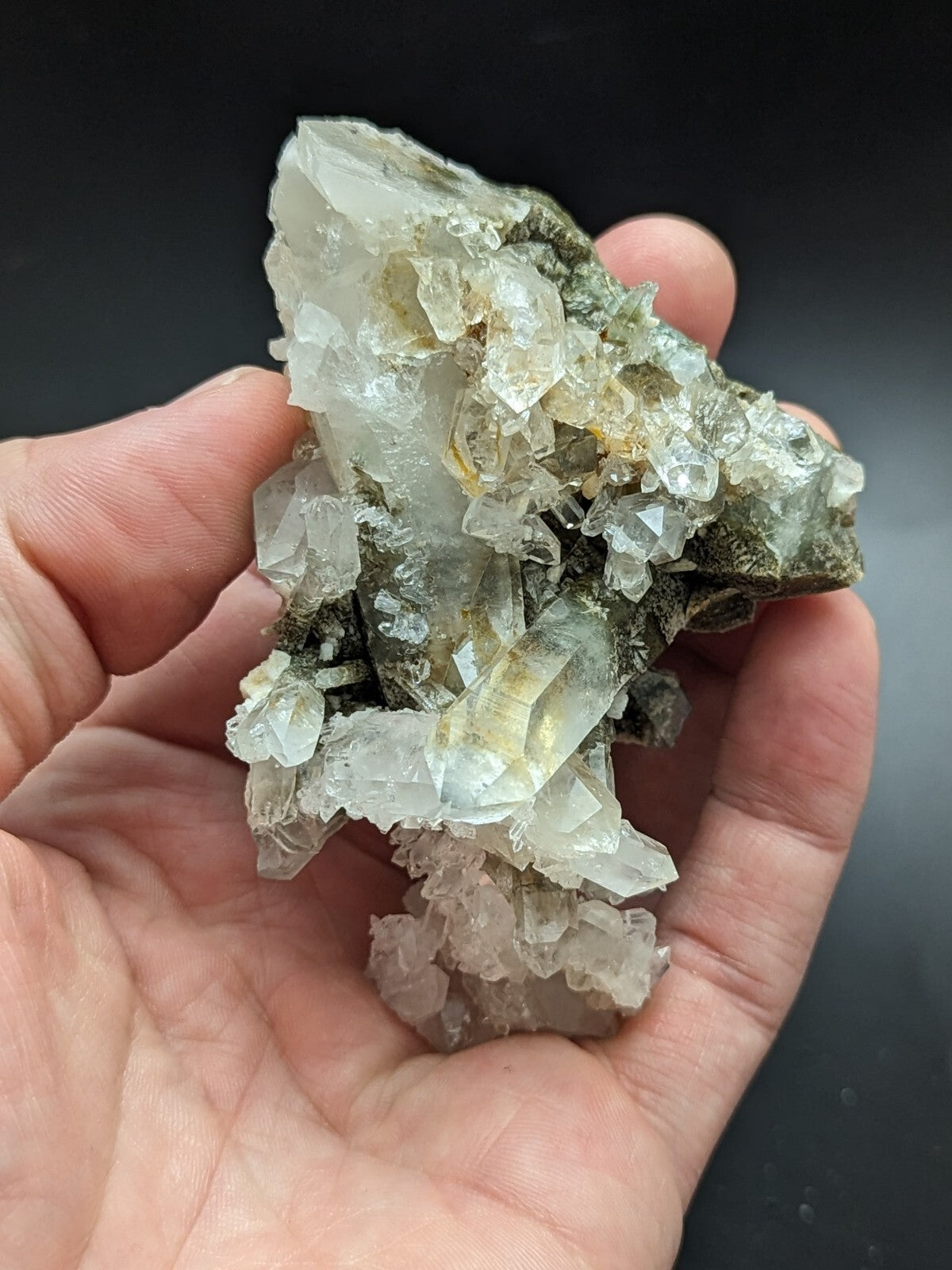 A hand holds a cluster of translucent, pale green and white quartz crystals growing from a dark mineral base.