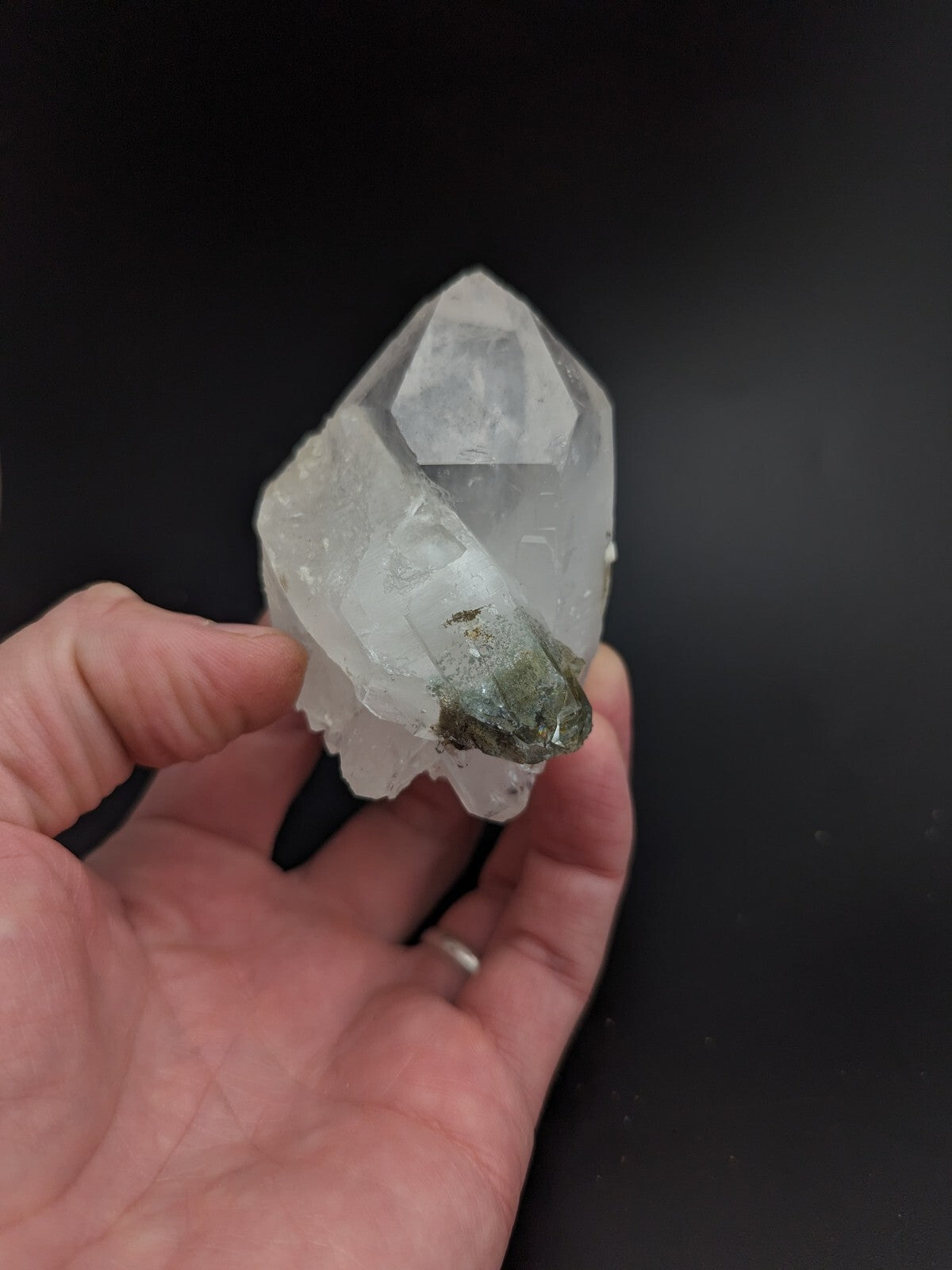A translucent white quartz crystal cluster with a greenish mineral inclusion at its base, held gently in someone’s hand.
