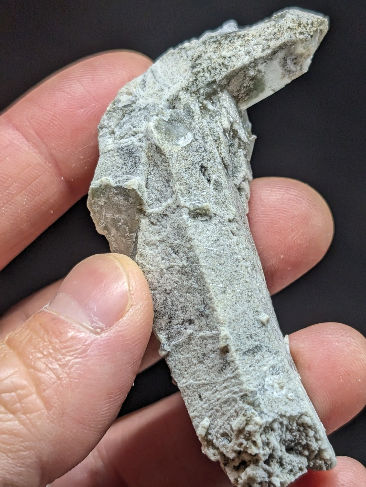 A rough, grayish-green mineral specimen with a jagged, irregular surface and visible crystalline formations.