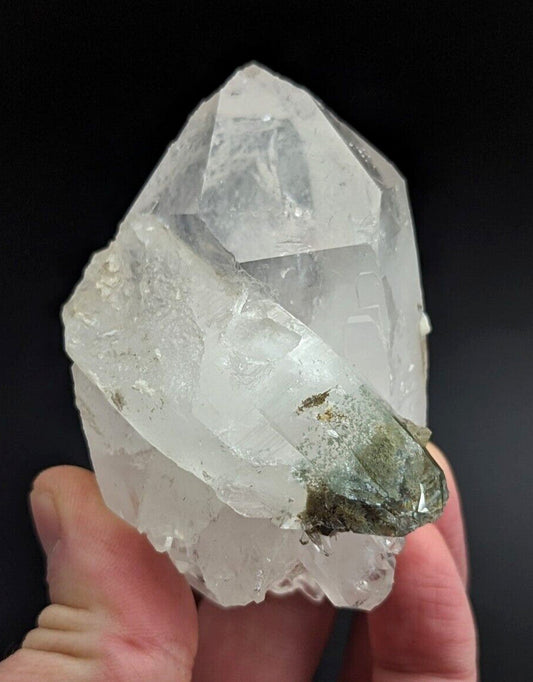 A translucent, clear quartz crystal cluster with a rough, faceted surface and a dark green mineral inclusion at its base.