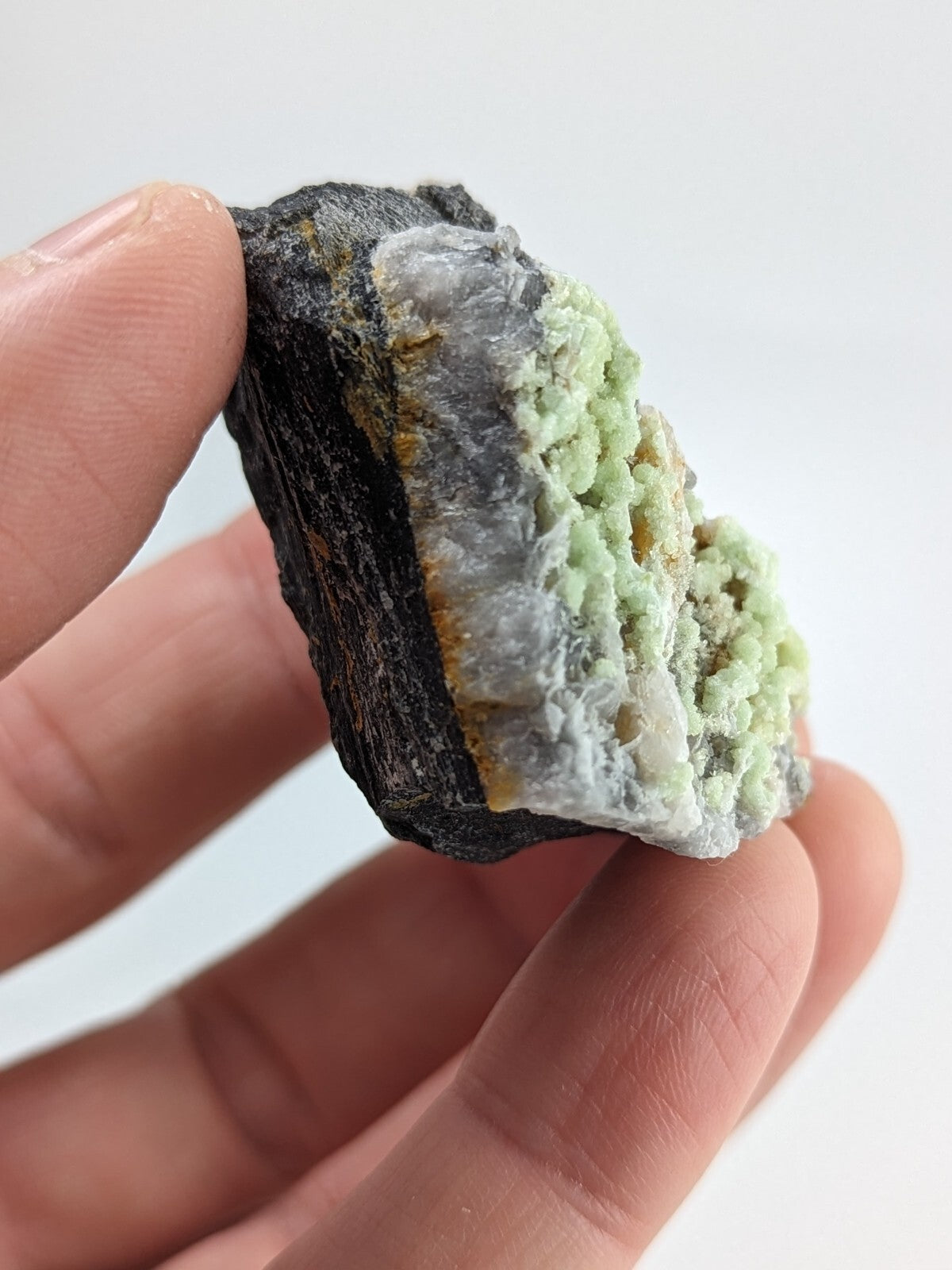 A hand holds a natural mineral specimen featuring a dark, rough-textured matrix with vibrant light green crystalline formations and subtle white veining.